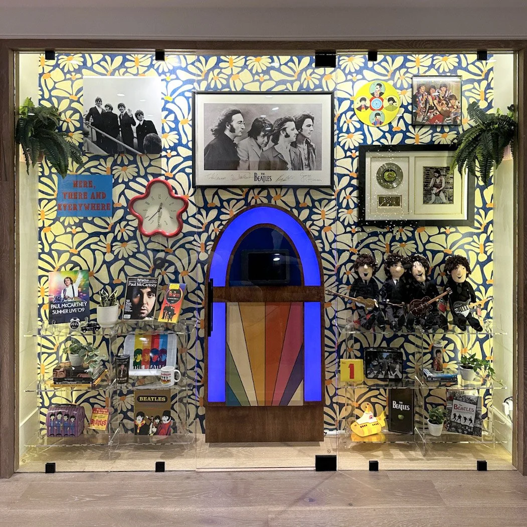 Decorated display wall featuring The Beatles memorabilia, photos, and artwork, with a rainbow-colored jukebox in the center and various potted plants.