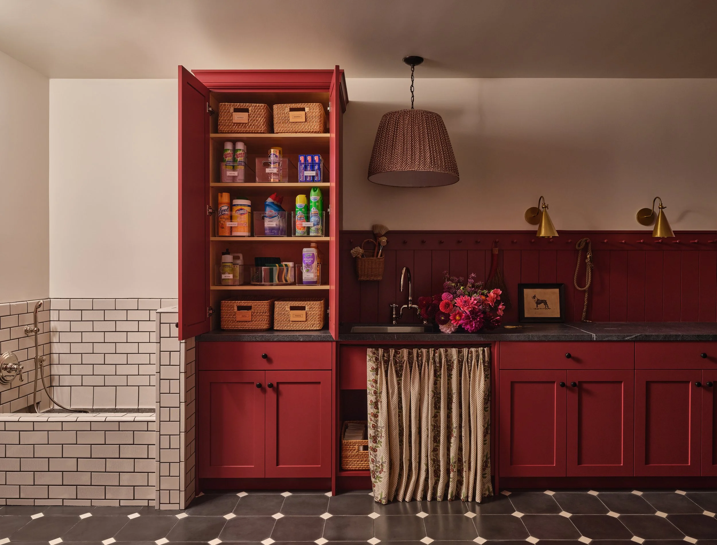 Kitchen with red cabinetry, open shelves holding woven baskets and cleaning products, floral curtains under the sink, black and white hexagonal tiled floor, wall-mounted lamps, pink floral arrangement.