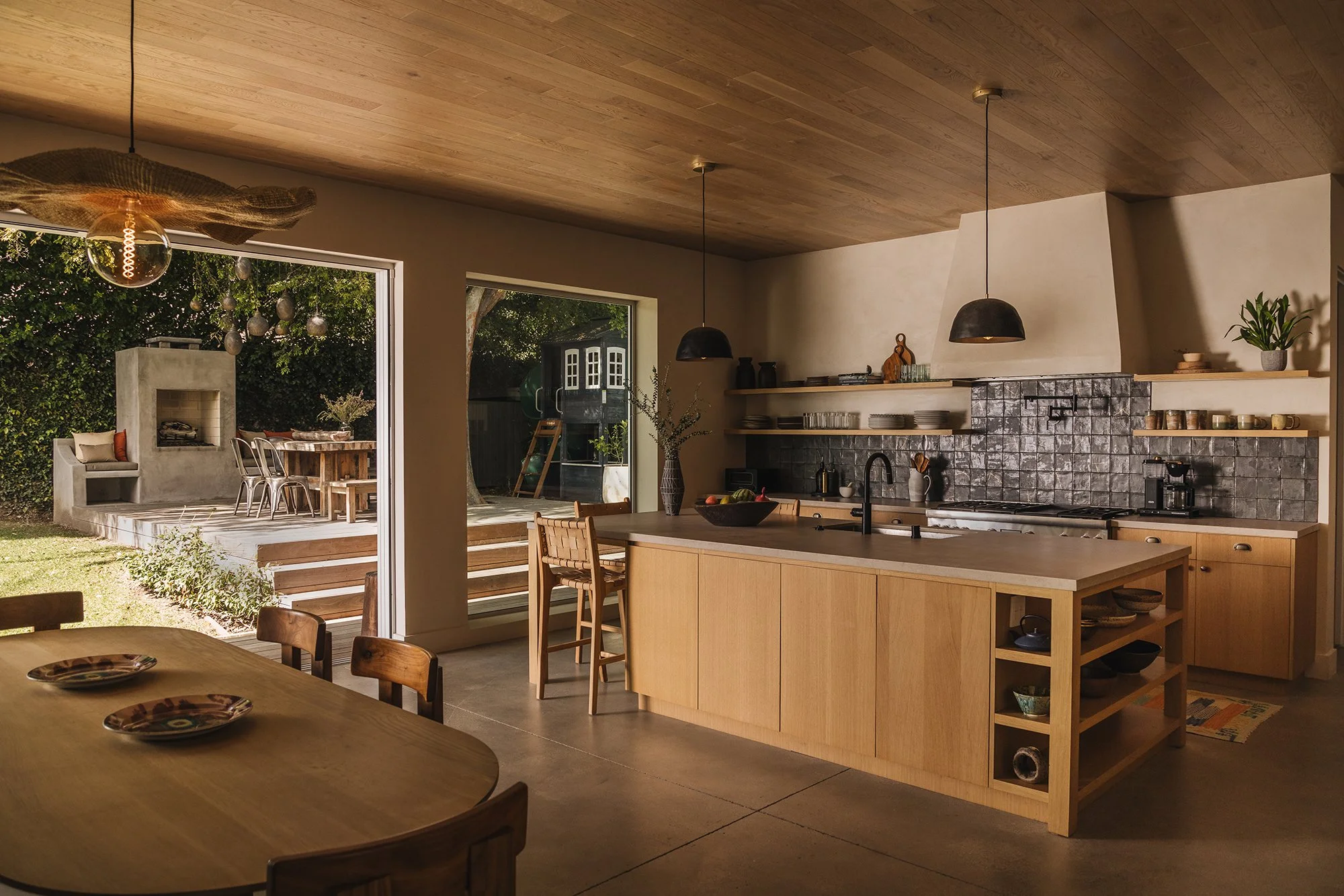 Modern kitchen with open doorway leading to outdoor patio and dining area, featuring trees and greenery.