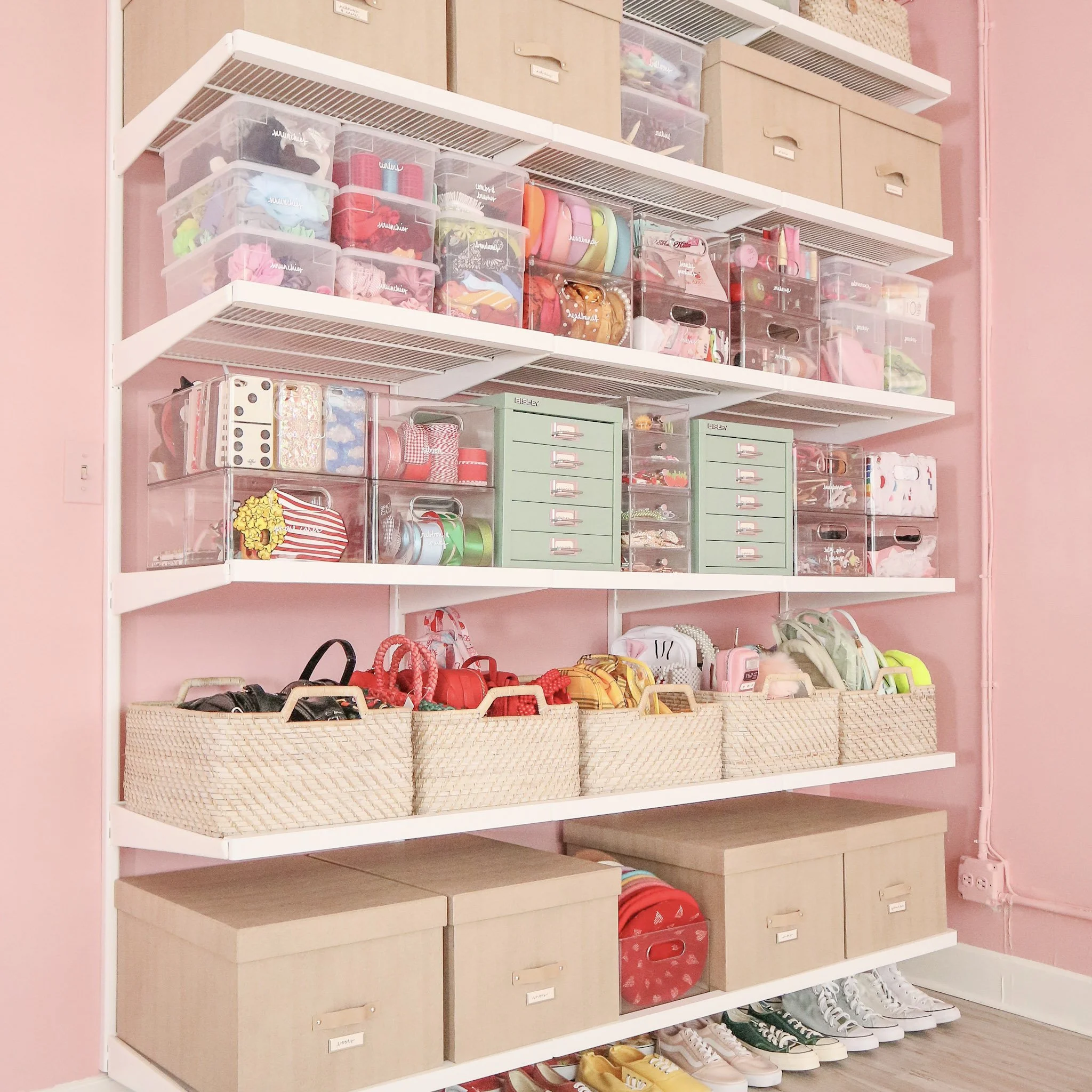 Open white shelving unit against pink wall, containing clear storage bins with shoes, accessories, makeup, and other items; beige storage boxes on top shelf; shoes organized on the bottom shelf.