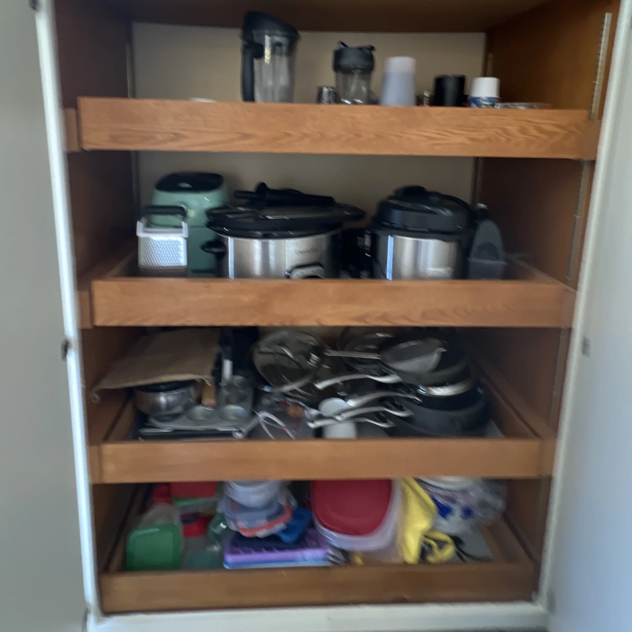 A wooden kitchen cabinet filled with various household items including small appliances, plastic containers, and other miscellaneous objects.