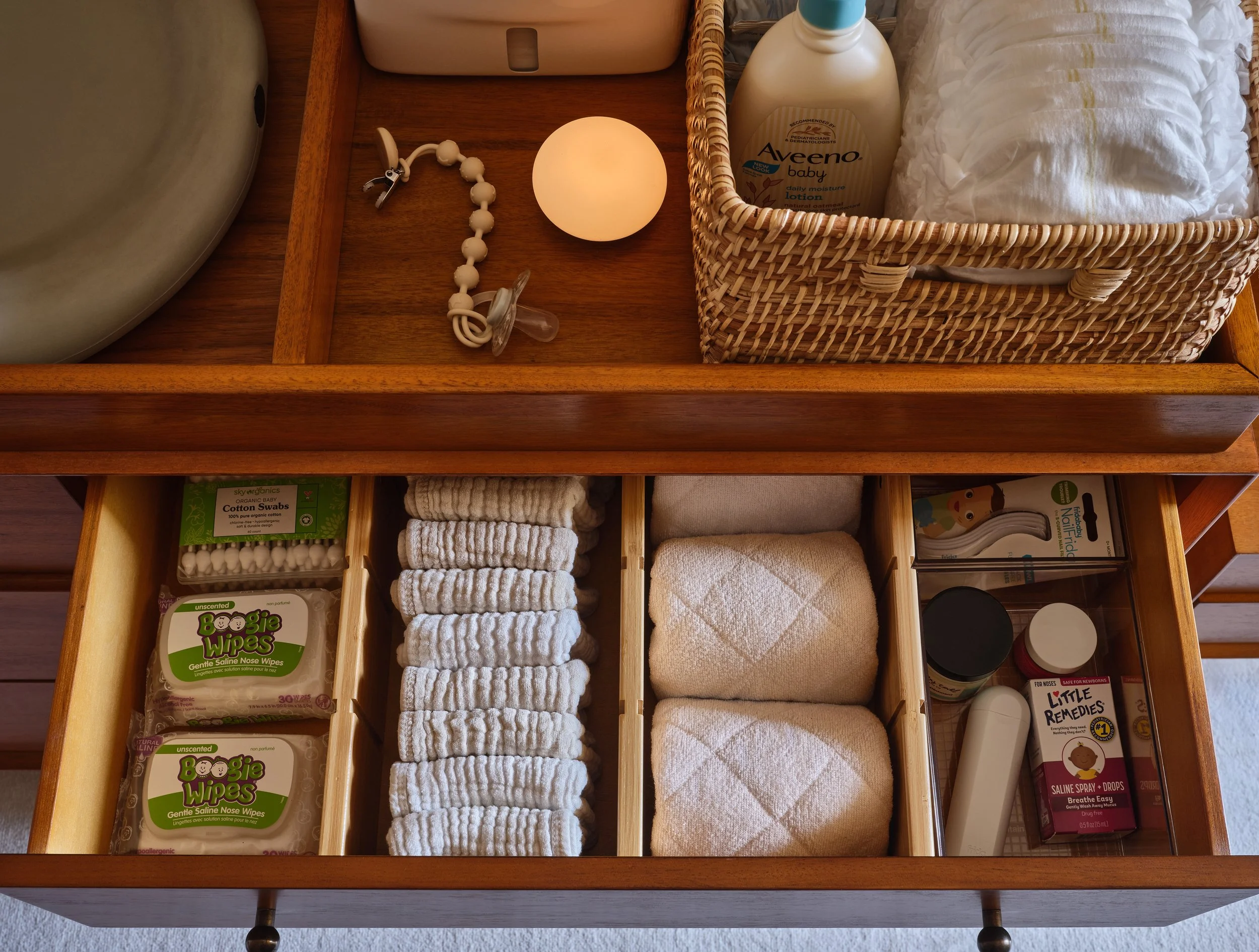 Open wooden drawer with baby items: white cloth diapers, baby lotion, pacifier, teething rings, and baby wipes, with some baby care products and a lamp on top.