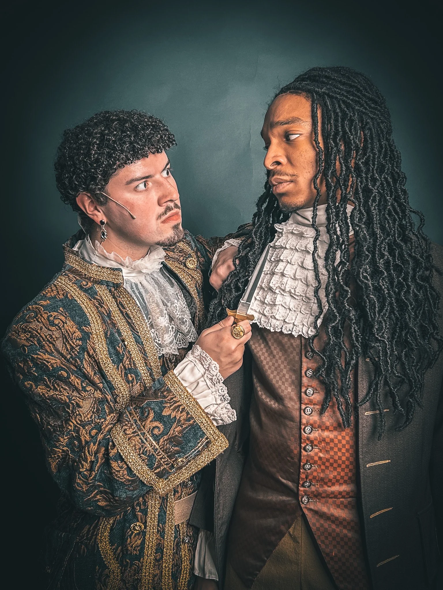 Two men dressed in elaborate historical costumes staring at each other intensely; one is holding a sword to the other's chest.