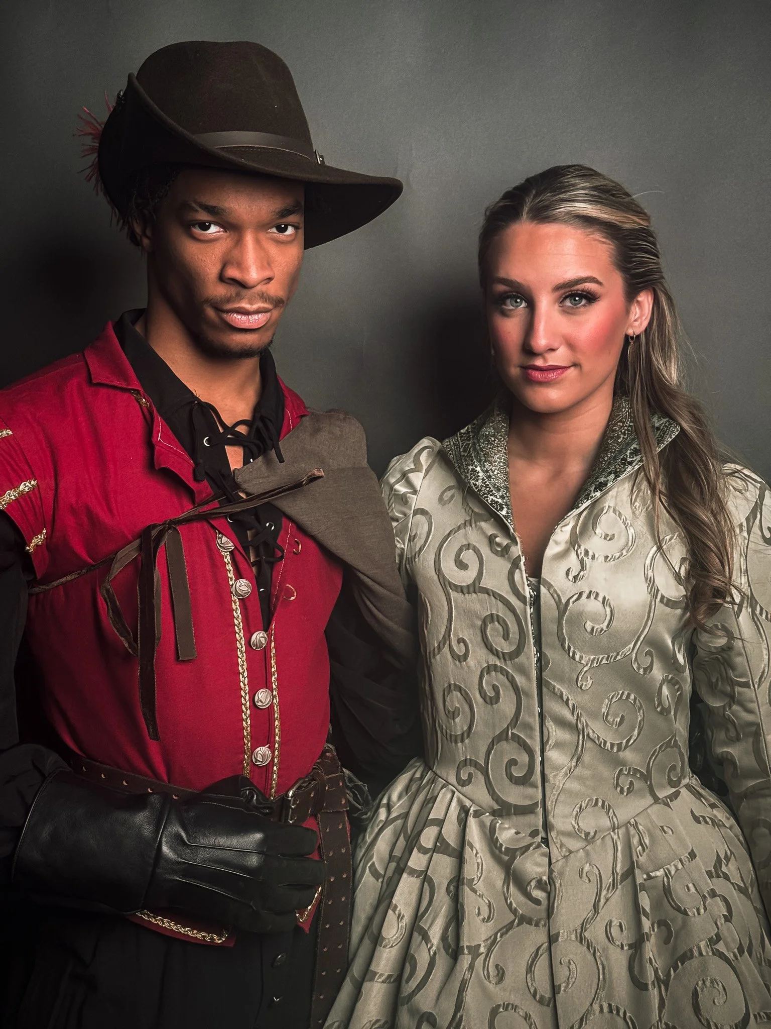 Two people dressed in costumes against a dark background, a man in a tattered red shirt with a wide-brimmed hat and gloves, and a woman in a patterned dress with a high collar and long sleeves.