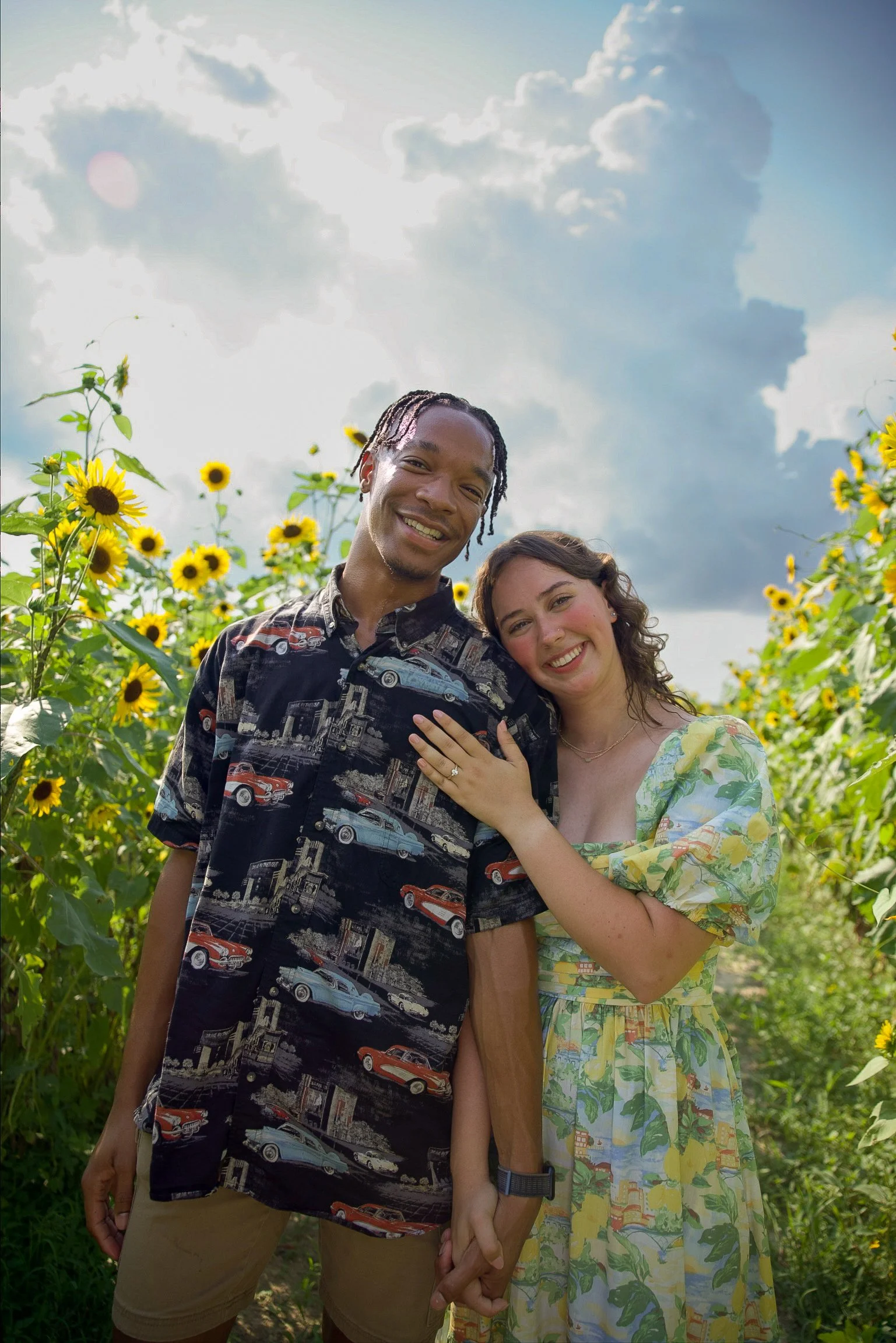 A smiling couple holding hands and embracing in a sunflower field under a partly cloudy sky.