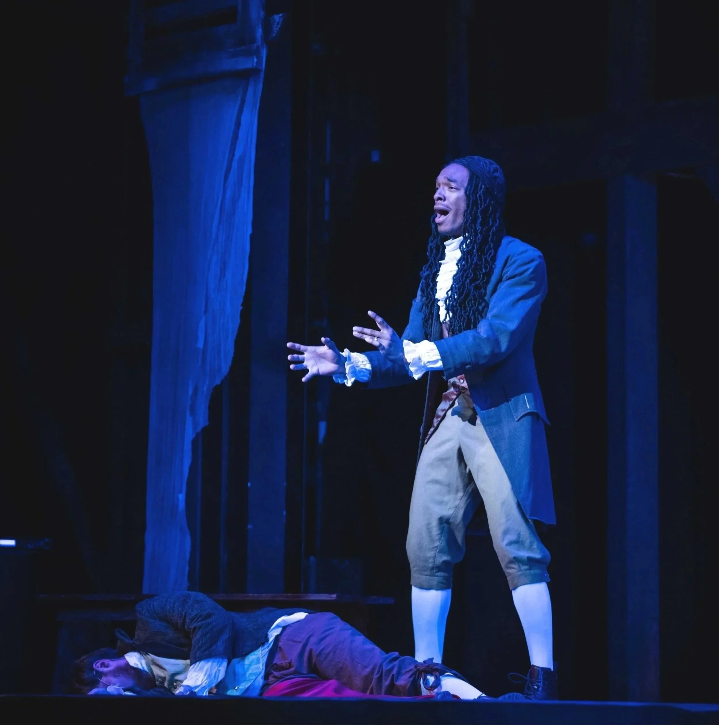 Justin Jackson is in A Tale of Two Cities,  playing Dr. Alexandre Manette, dressed in historical costume singing passionately on stage, with a person lying on the floor in period clothing in front of him, and a dark background with stage props.