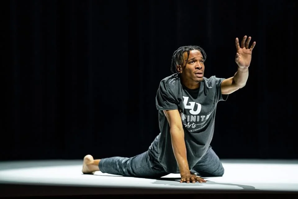 Justin Jackson is seen with braided hair performing a theatrical or dance pose on a stage, reaching forward with one arm and supporting himself with the other, against a dark background.