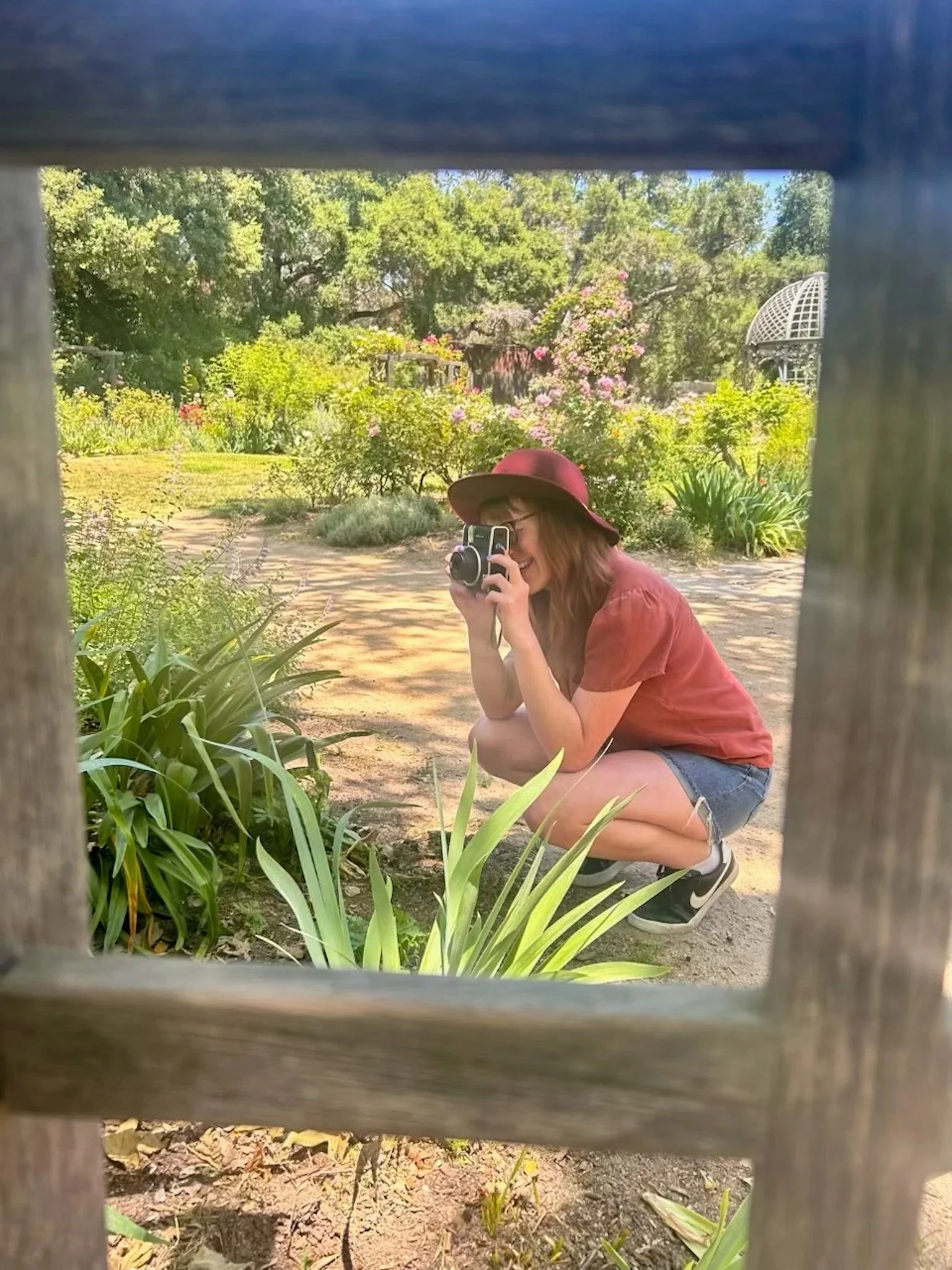 A photo of Lauren outside among plants, crouched in an orange t-shirt and jean shorts, holding a camera and snapping a photo.
