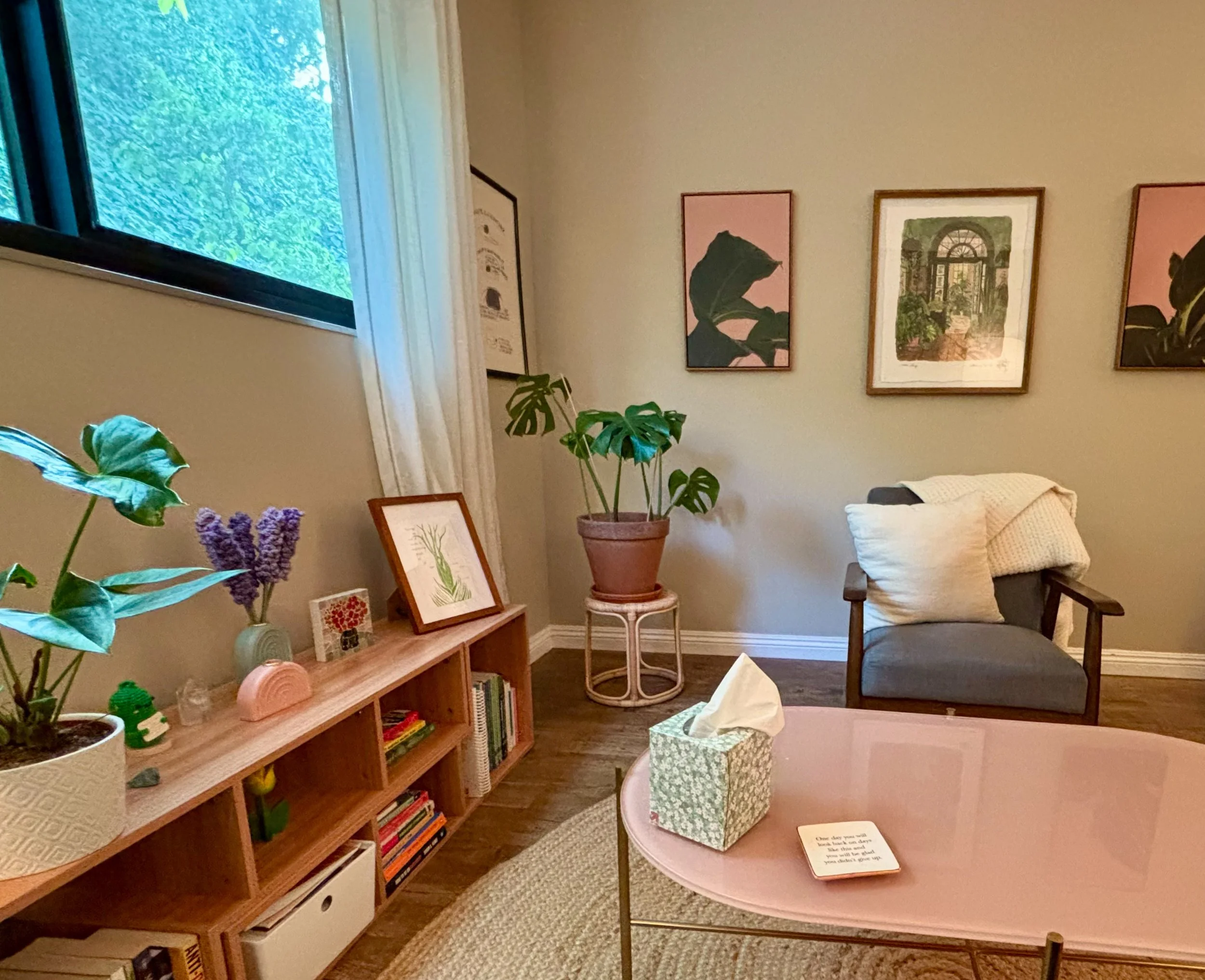 A photo of Lauren's office. On the left there is a horizontal shelf full of books. On top of it are plants, and various pieces of art. In the middle of the room there is a pink coffee table and a gray chair.