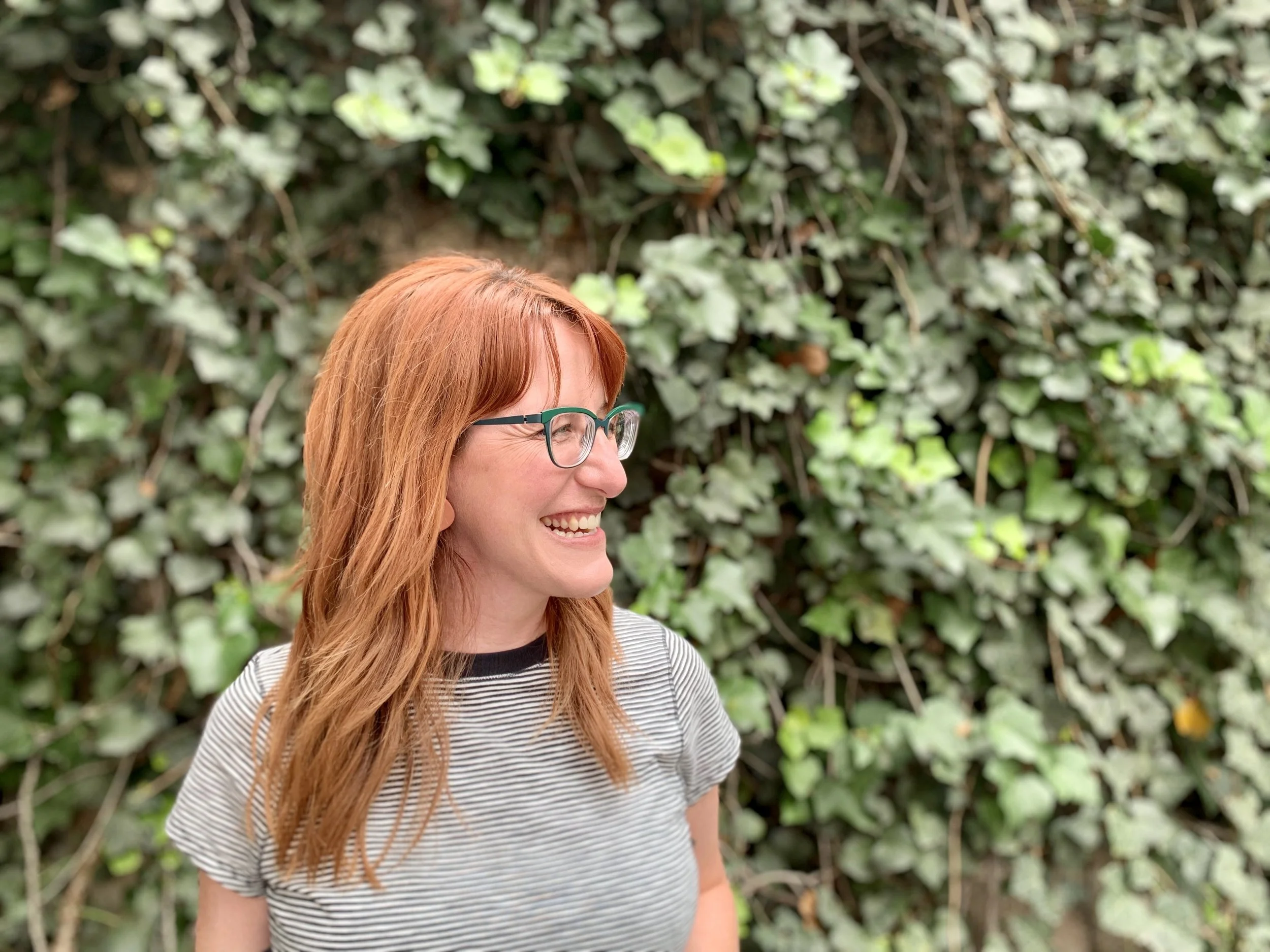Lauren (a white woman with long red hair) wears a black and white striped t-shirt and emerald green glasses. She's smiling and standing in front of a wall covered in green ivy.