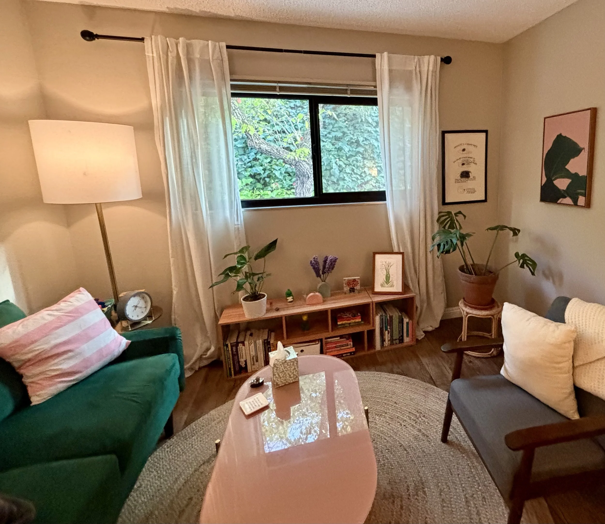 A photo of Lauren's office. There is an emerald couch and gray chair, pink coffee table, horizontal bookshelf, and lots of plants and art.