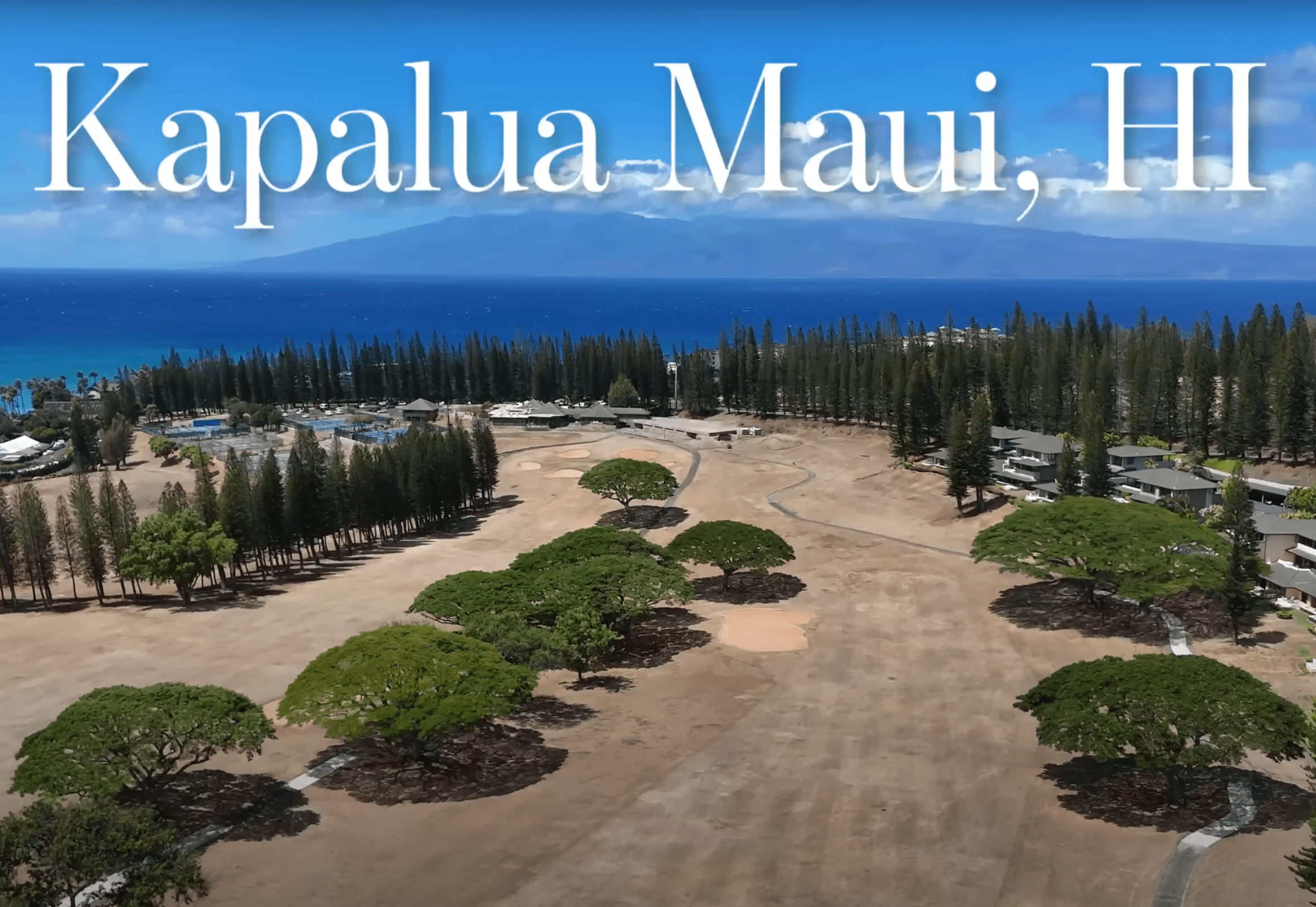 Aerial view of Kapalua, Maui, Hawaii, showing a dry and brown golf course, trees, houses, and the ocean with mountains in the background.