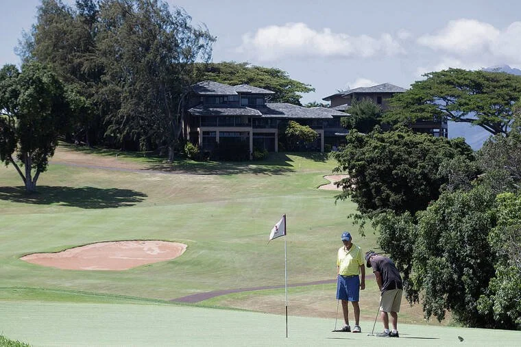 Two men playing golf on a course with a building in the background and trees surrounding the area.
