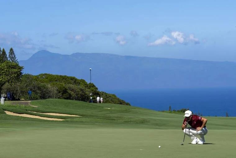 A golfer crouching on a golf course preparing for a putt with ocean and mountains in the background.