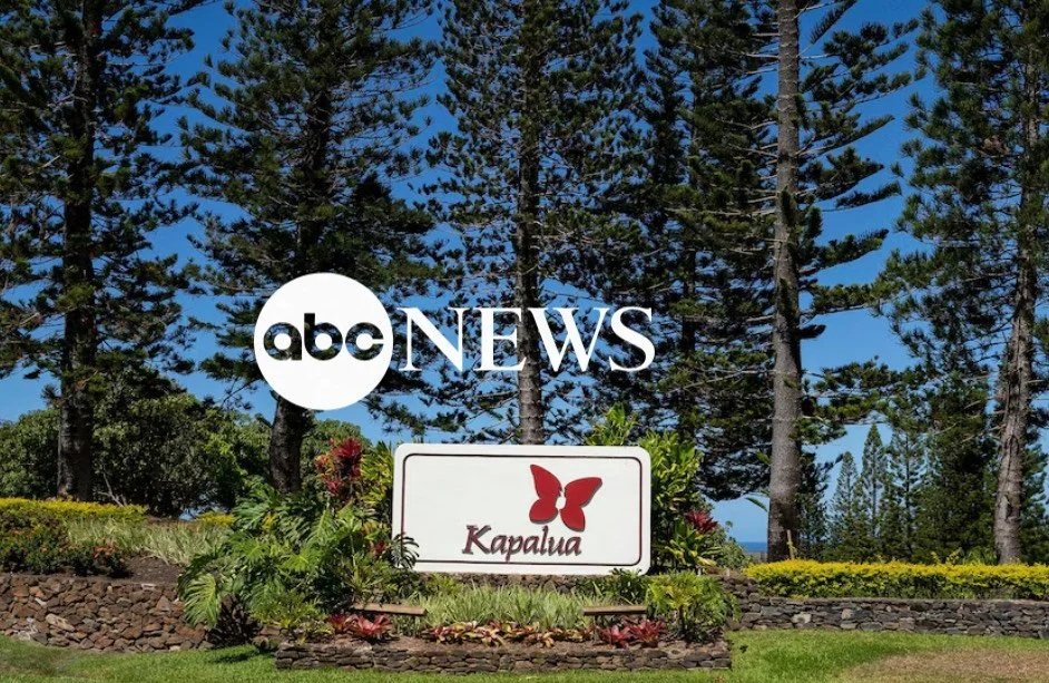 A landscaped area with a sign that reads "Kapalua" and a butterfly logo, surrounded by trees and greenery, under a blue sky.