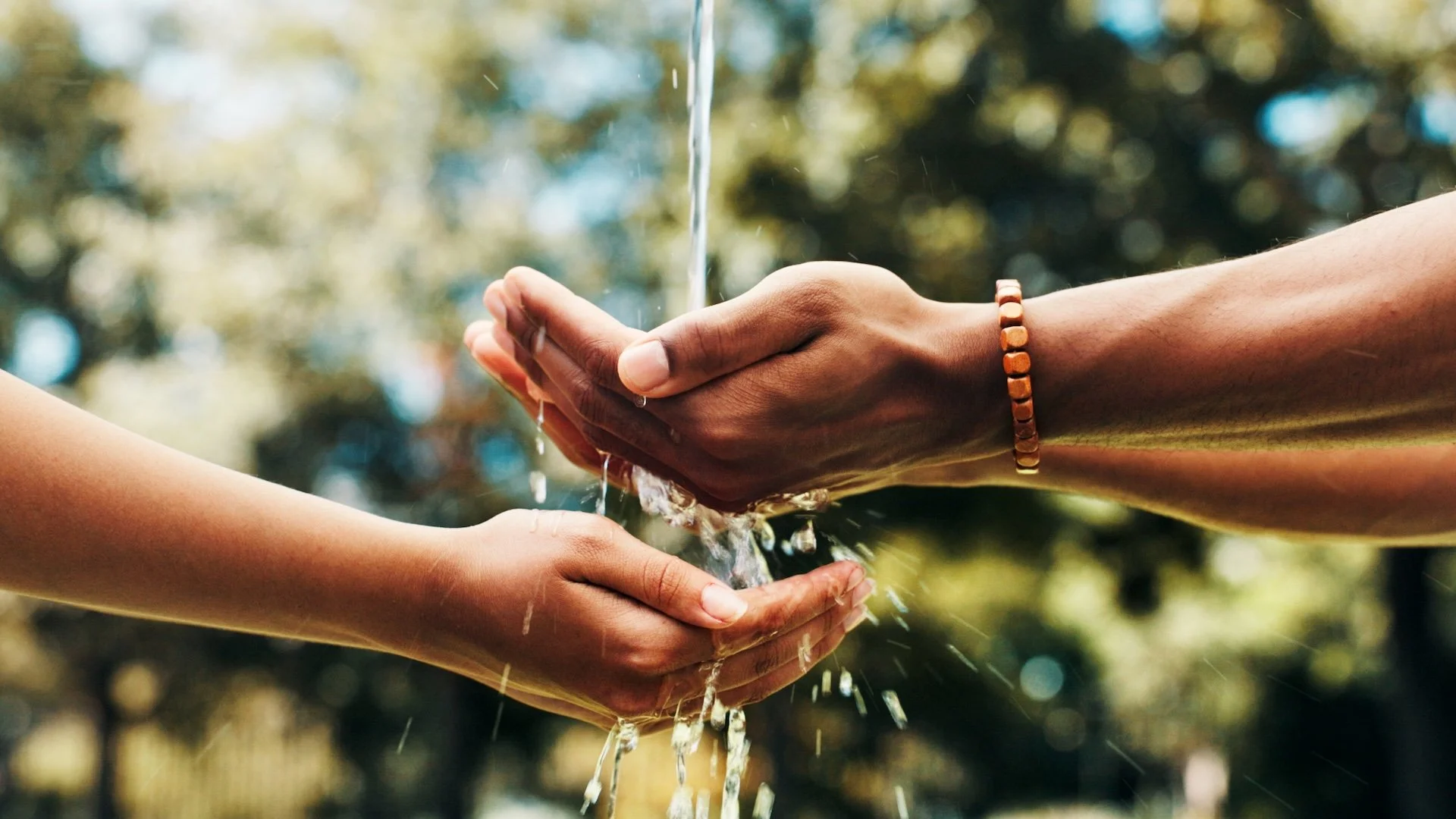 Two people holding hands while washing each other's hands with water outdoors, blurred background with trees.