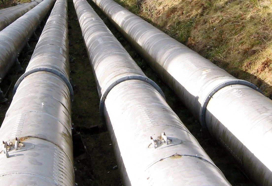 Four large metal pipelines lying on the ground with stands securing them, in an outdoor setting with grass on the side.