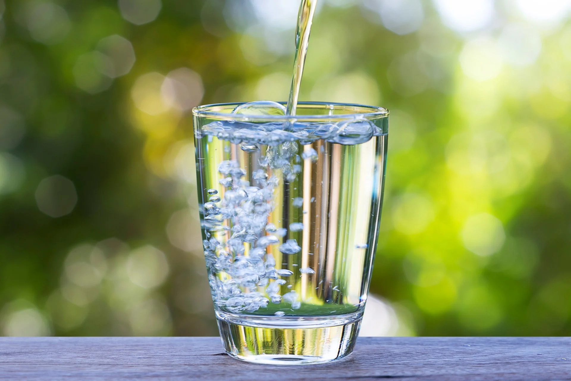A glass of water being filled from above, with bubbles forming as water flows into the glass, against a blurred green and yellow background.