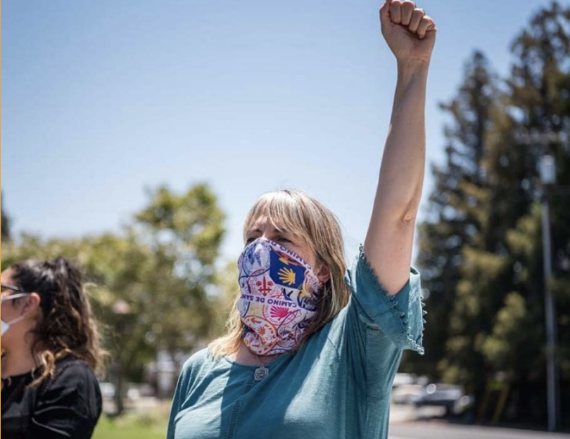 A woman wearing a colorful mask with a yellow hand logo and the words "Land of the Free" raises her fist in a show of protest or solidarity outdoors on a sunny day, with trees in the background.