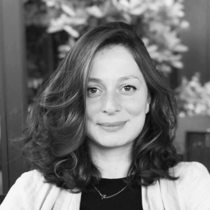 Black and white portrait of a woman with wavy shoulder-length hair, smiling softly, with a blurry background of plants or flowers.