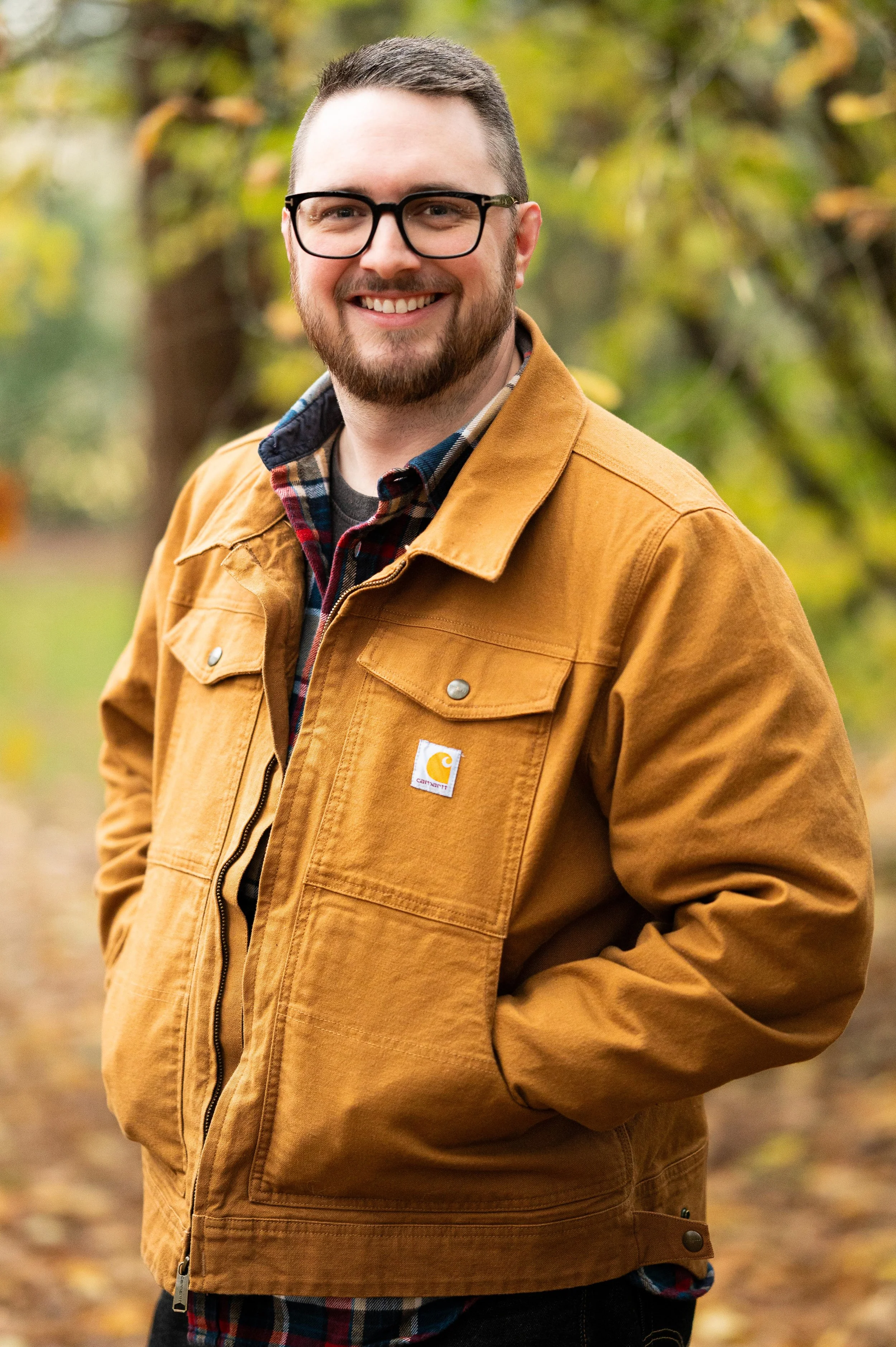 A smiling young man with glasses and a beard standing outdoors in a park with fall trees in the background, wearing a tan Carhartt jacket over a plaid shirt.