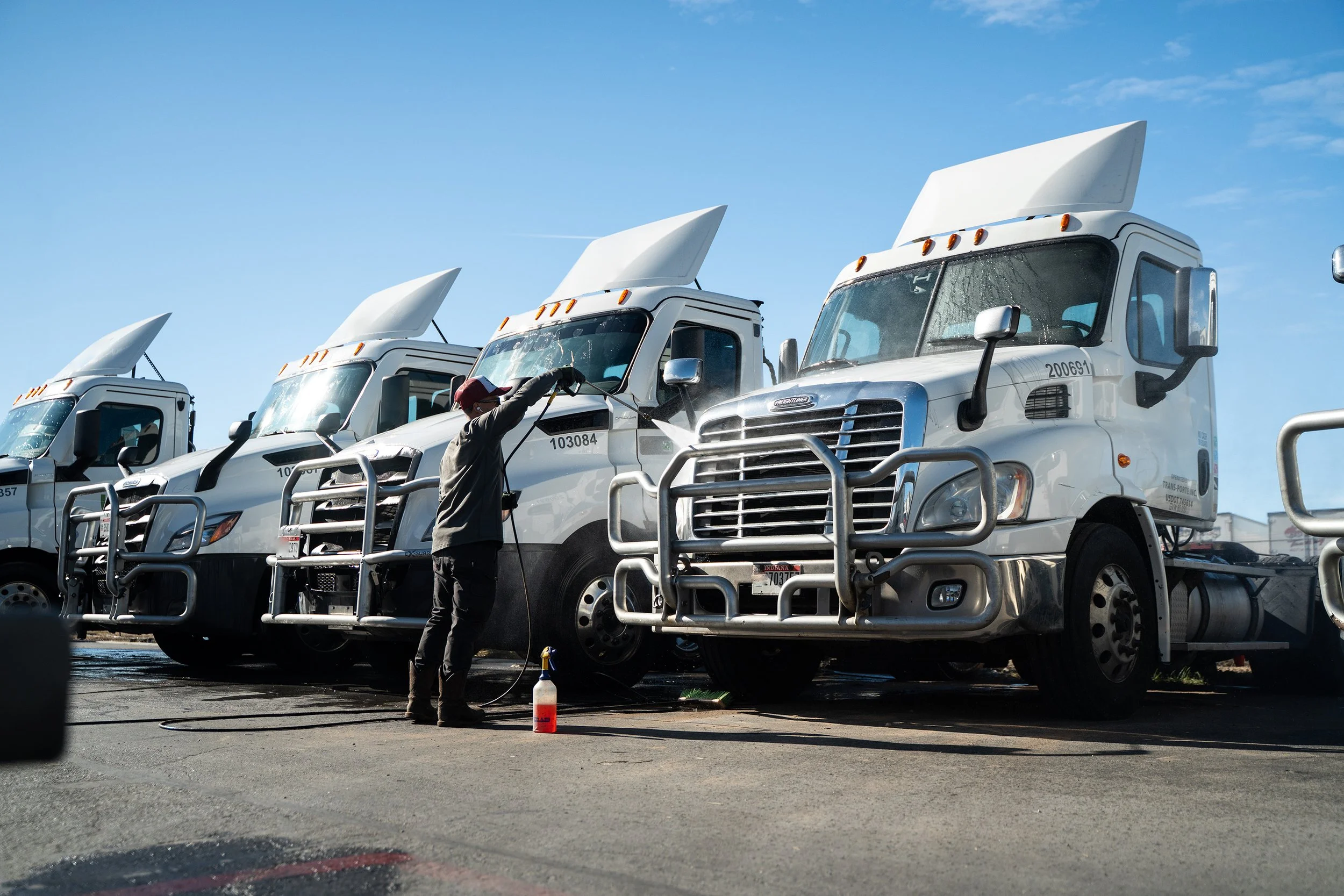 A person cleaning or washing large semi-truck cabs parked in a row outdoors on a sunny day.