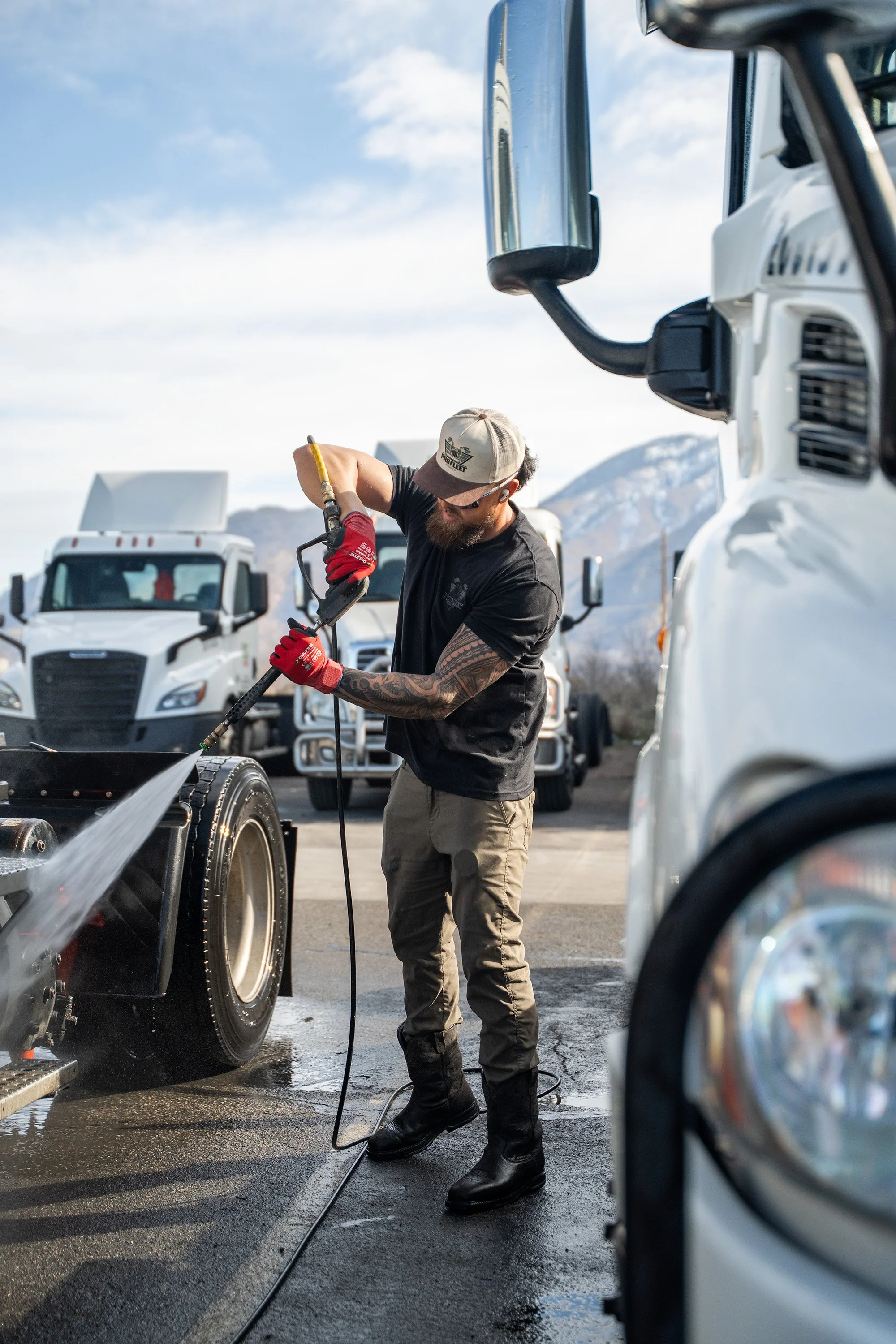 Man washing a large truck with a high-pressure hose outdoors, with other trucks in the background.