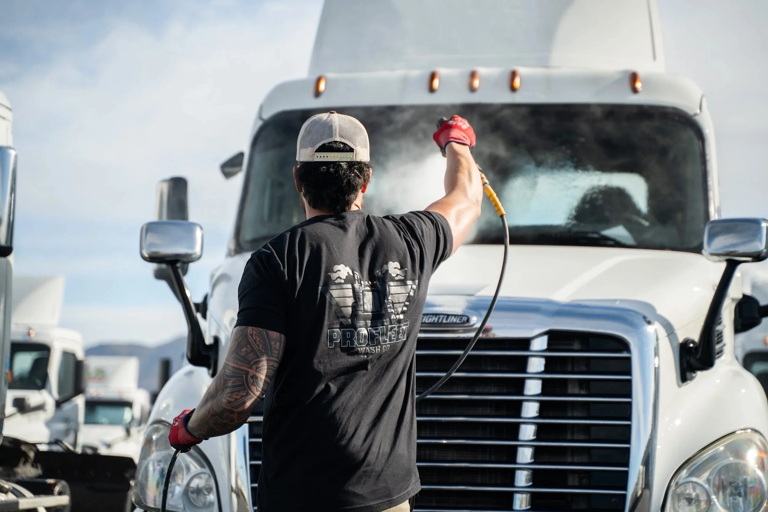 A person wearing a black T-shirt, a gray cap, and red gloves is pressure washing the front windshield of a large white truck. The person has a tattoo sleeve on the right arm. The scene appears to be outdoors on a sunny day with other trucks in the background.