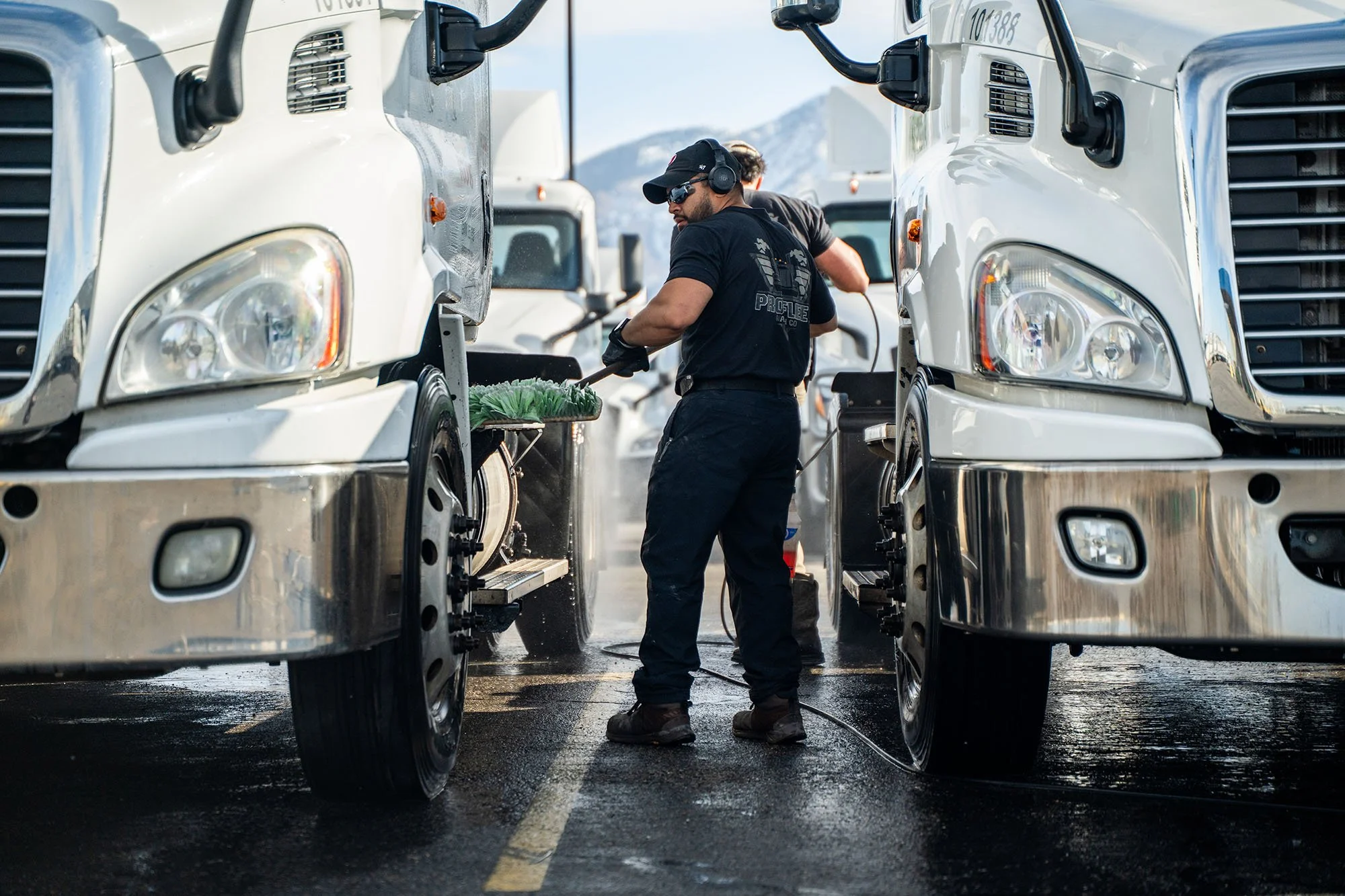 A truck washer wearing black clothing, gloves, and a cap, is cleaning the wheels of a white semi-truck in an outdoor lot while wearing noise-canceling headphones, with other trucks visible in the background.