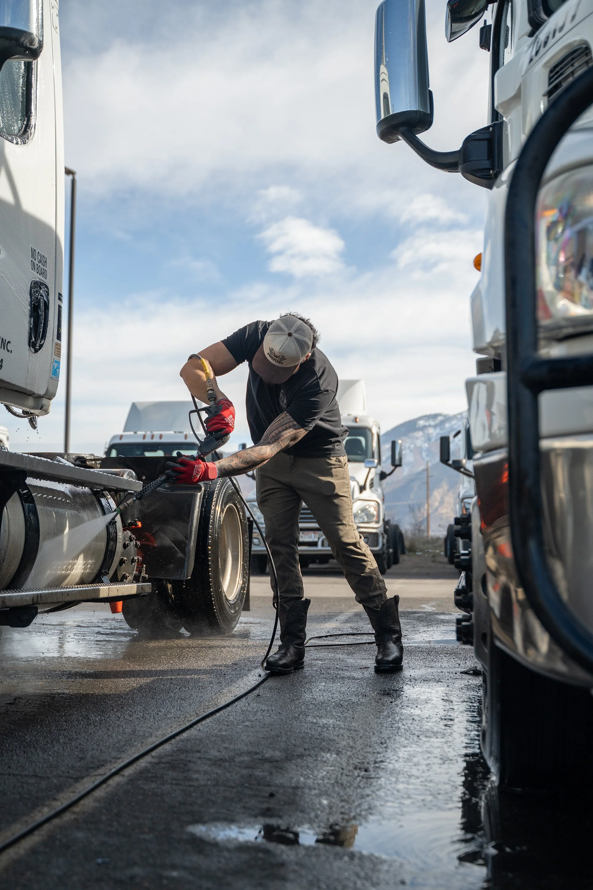 A man wearing a gray cap, black t-shirt, khaki pants, and black boots is pressure washing the wheel of a large commercial truck, with other trucks visible in the background and mountains under a partly cloudy sky.