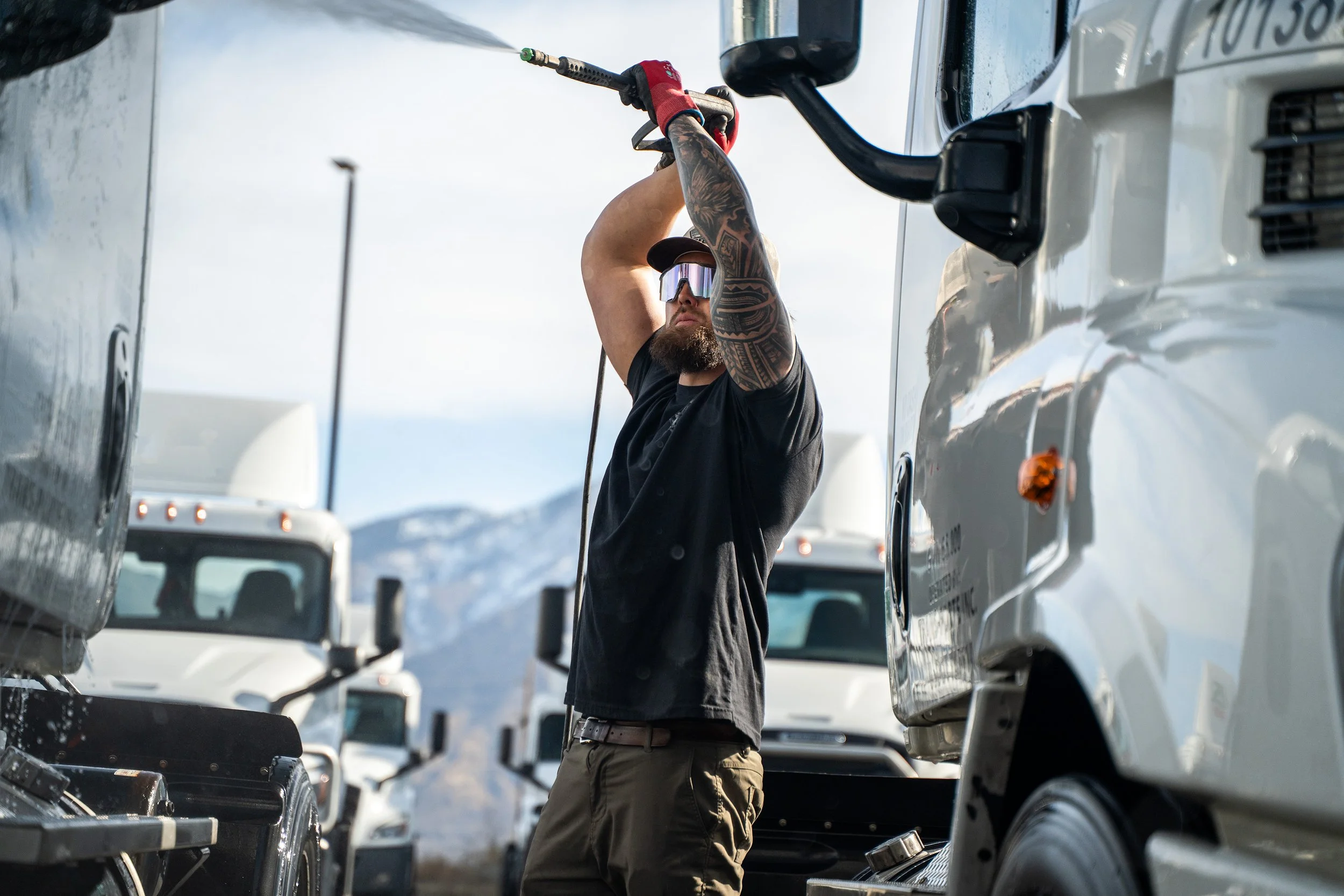 A man with tattoos, wearing a black shirt, sunglasses, and a cap, is washing a large white truck with a high-pressure hose outdoors near a snowy mountain range.