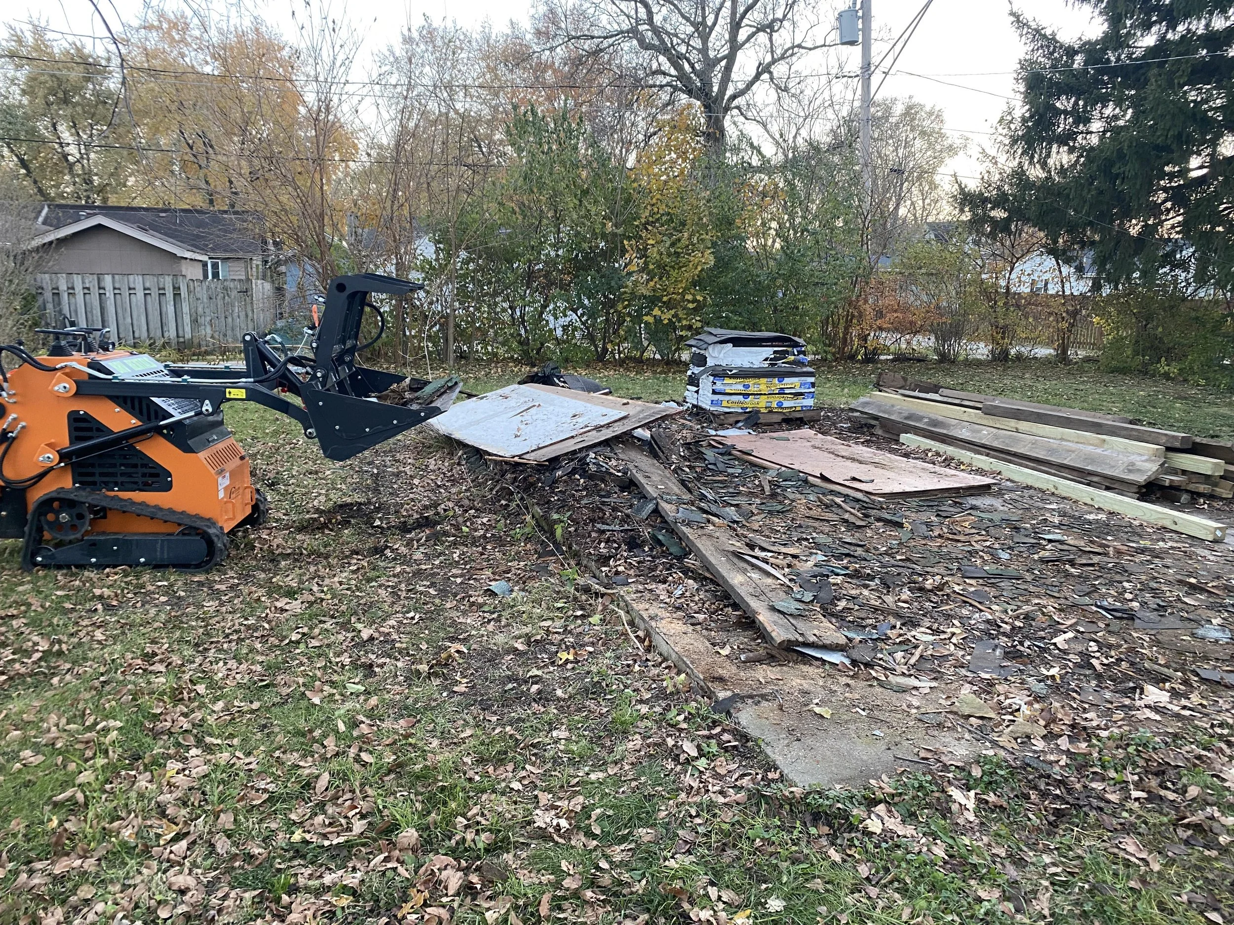 A backyard scene with construction materials and equipment. There is a small orange and black power tool or machine on the left, some wooden planks and boards on the ground, and several bags of insulation stacked near a bush. Junk removal sevice.