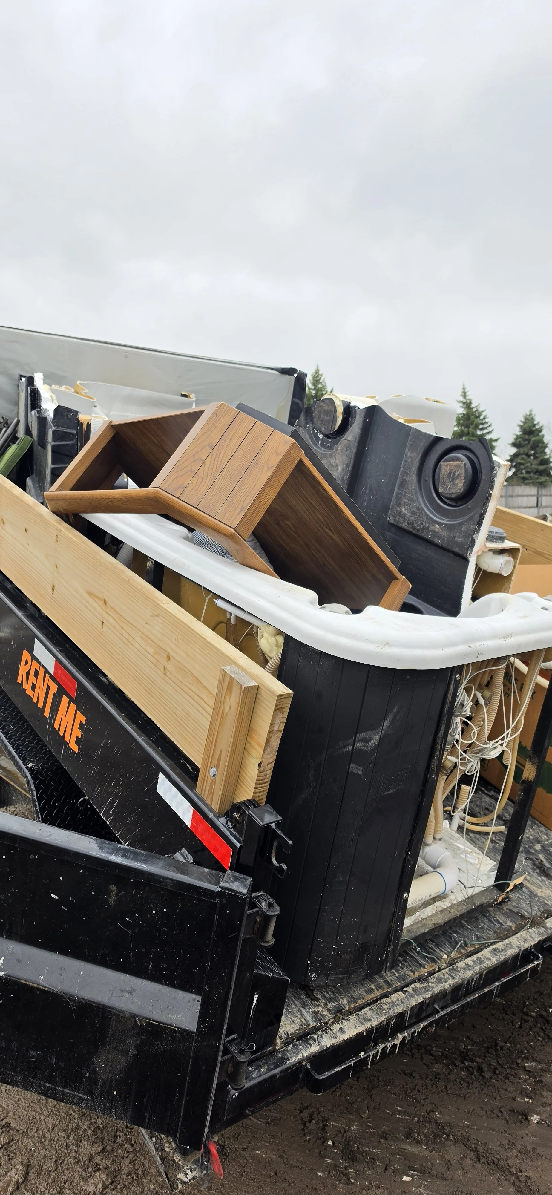 Trailer filled with household items including a wooden shelf, speakers, broken appliance parts, and tubing on a muddy ground under an overcast sky. Junk Removal Service.