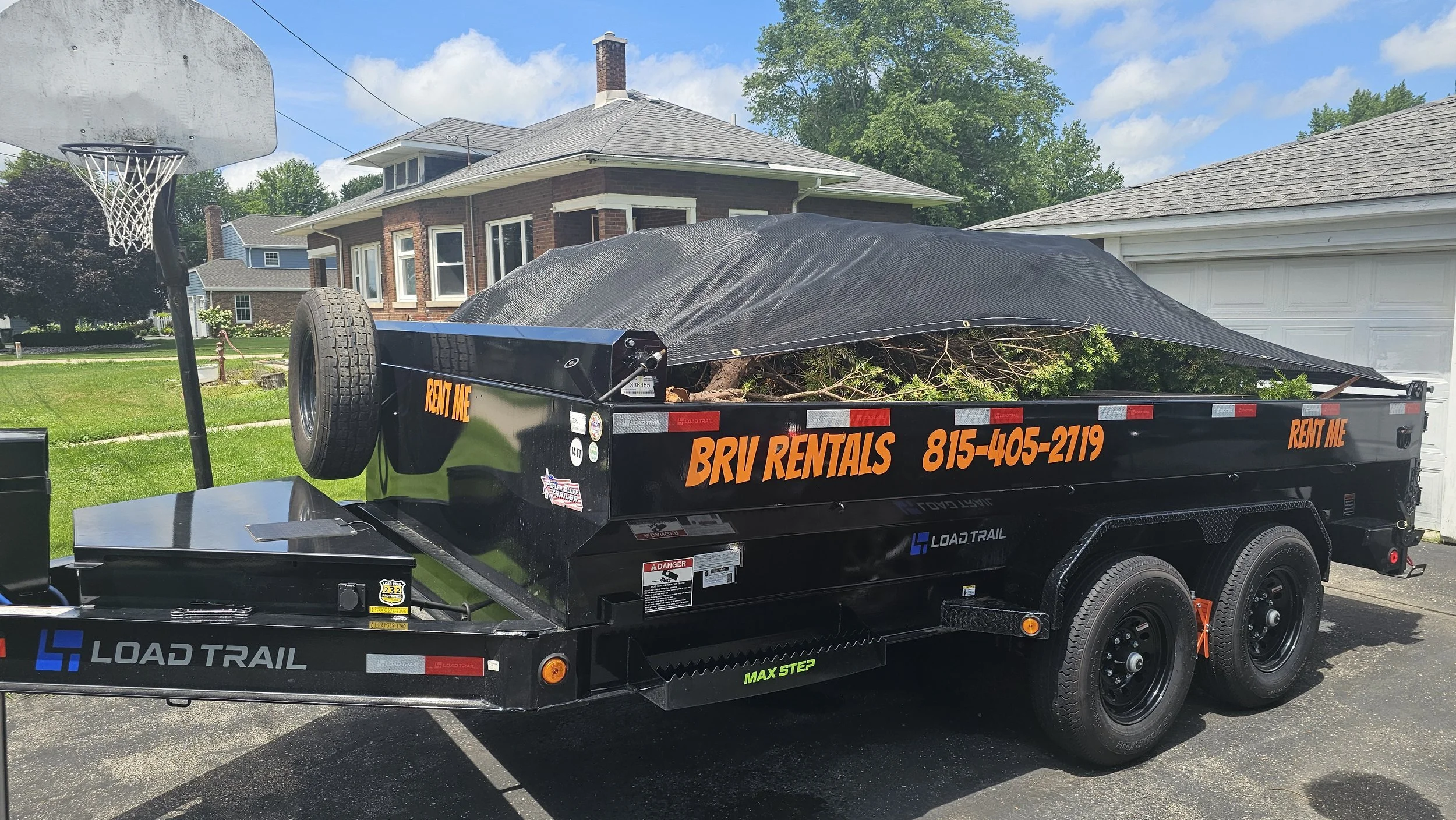 A black flatbed trailer loaded with yard debris parked in a driveway in front of a house. The trailer has orange text advertising BRV Rentals with a phone number. Junk Removal service, dumpster and dump trailer rental.