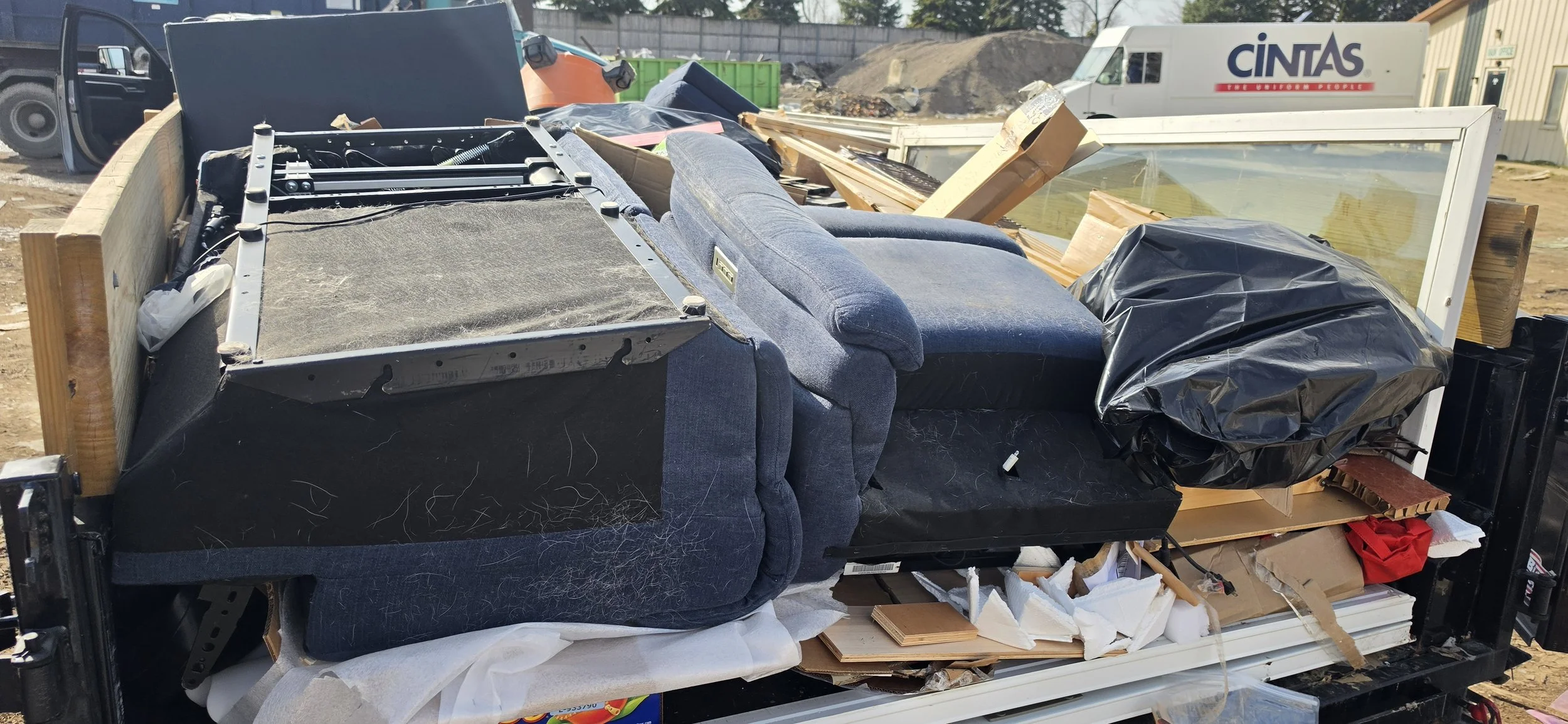 A large truck or trailer filled with various discarded furniture and debris, including a sofa, window, and other miscellaneous items, parked in a dirt lot. Junk Removal service and dumpster rental.