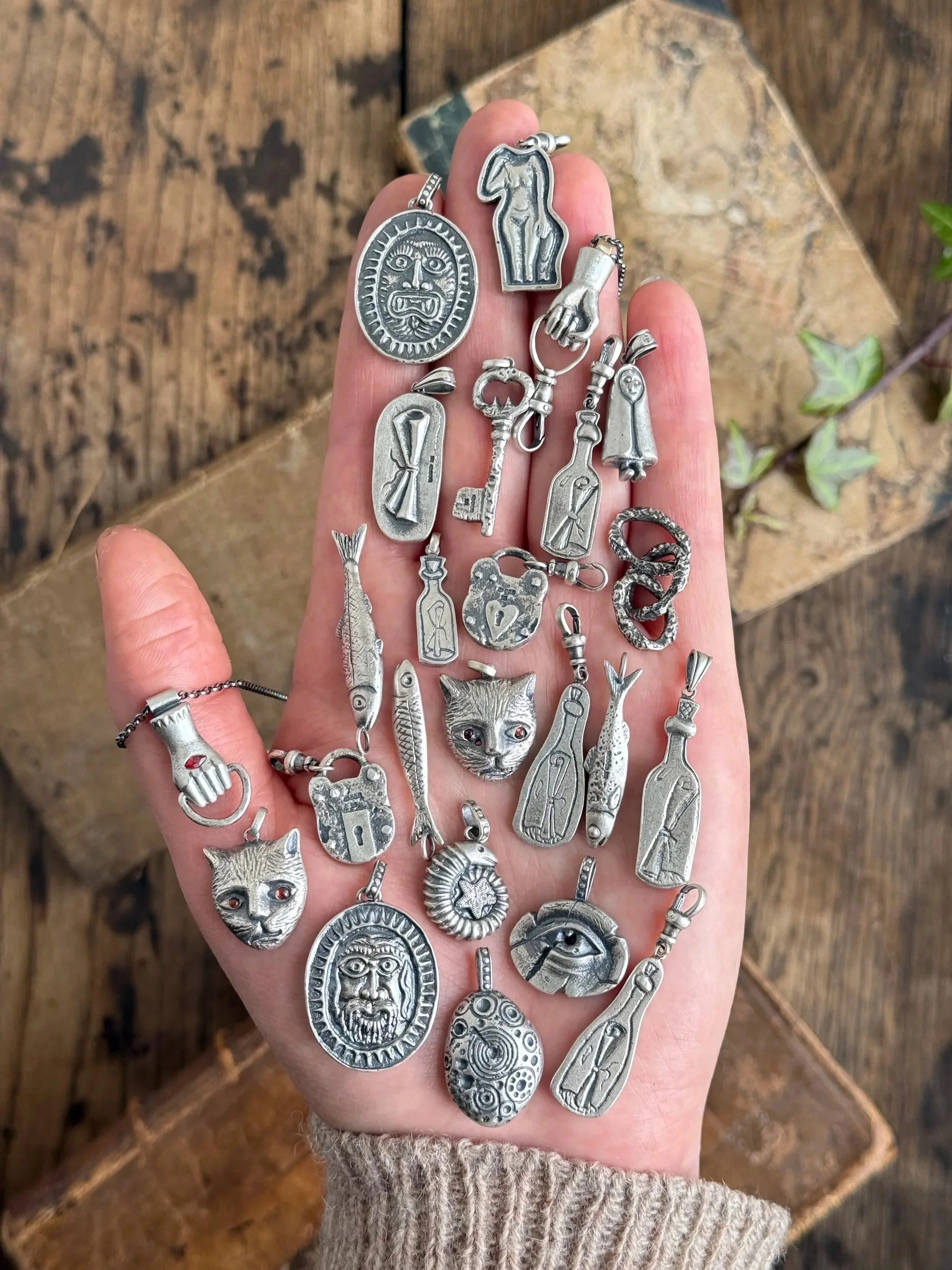A person's hand holding a variety of silver jewelry charms, including animal faces, keys, bottles, and abstract shapes, on a wooden surface.
