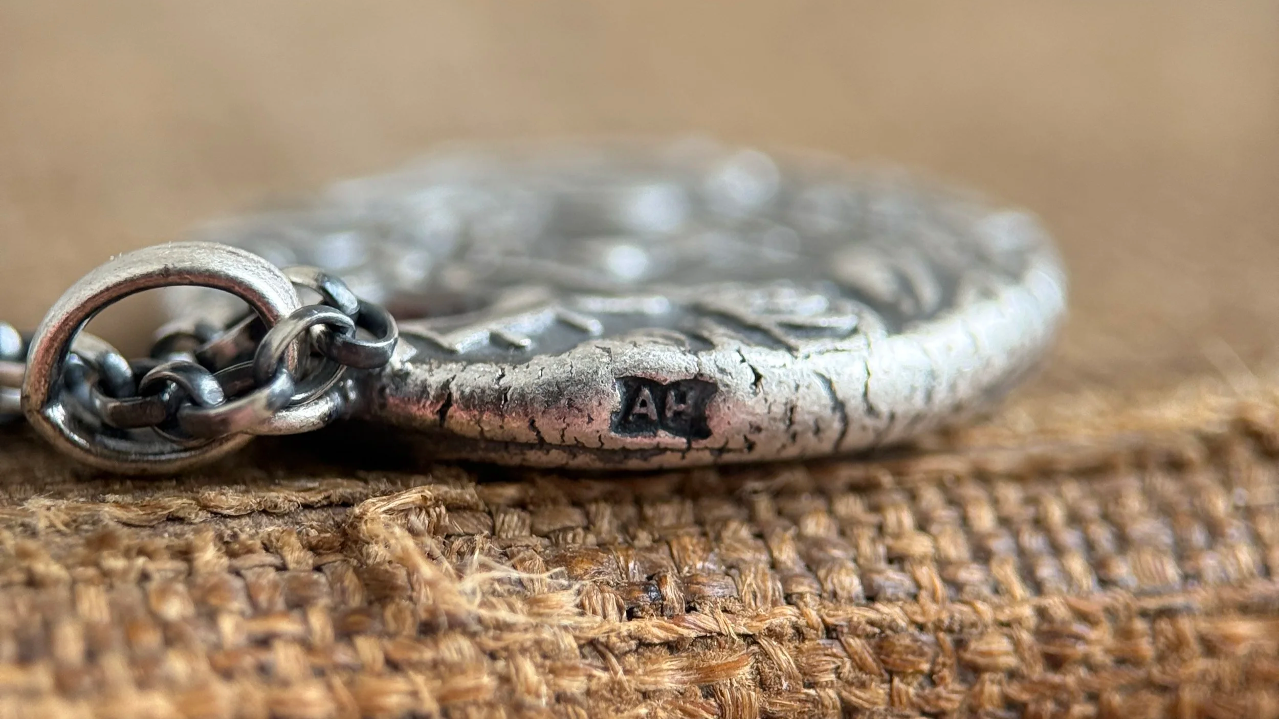 Close-up of a silver metal pendant with an insignia, attached to a chain, resting on a woven brown fabric surface.