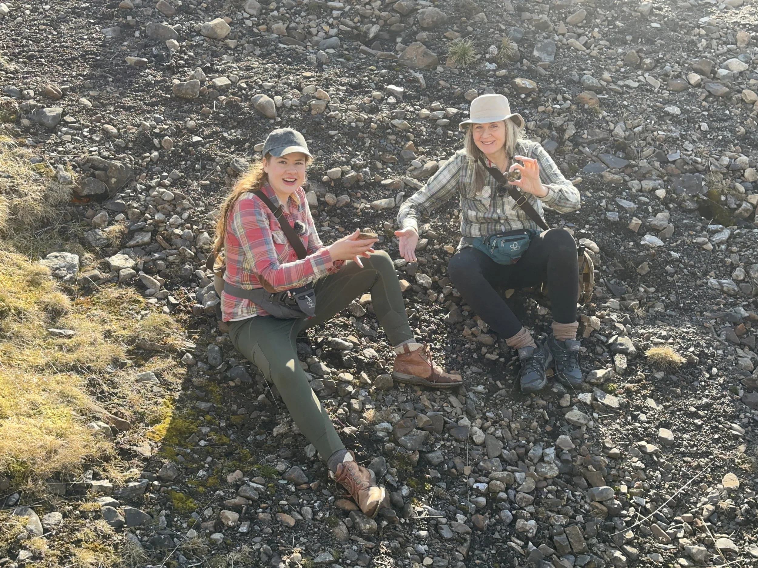 Two women sitting on rocky ground outdoors, smiling while holding crystals, wearing hiking gear and plaid shirts.