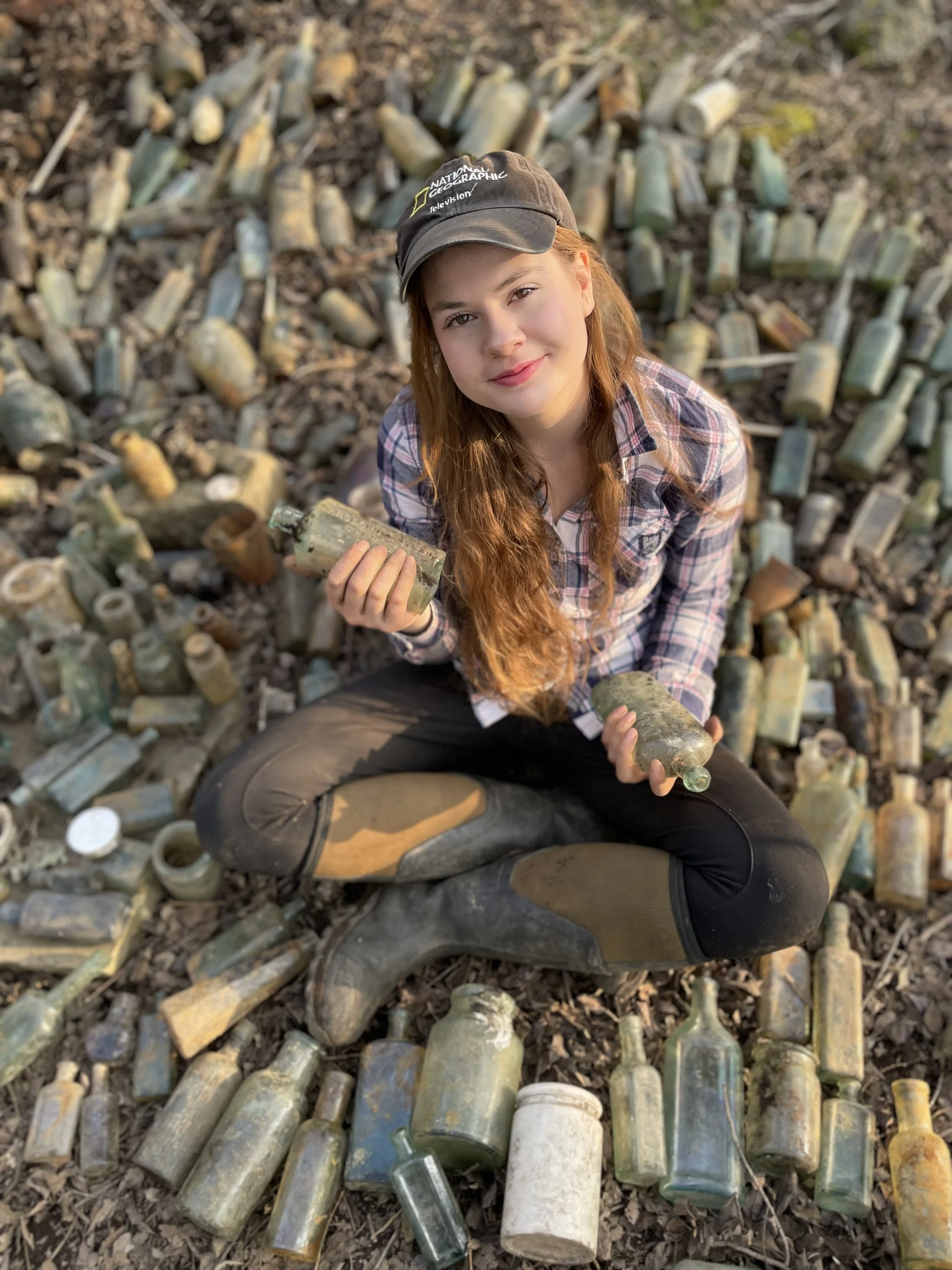 A young woman with long red hair, wearing a plaid shirt, black pants, rain boots, and a baseball cap, sits on the ground surrounded by broken glass bottles and holds two glass bottles in her hands.