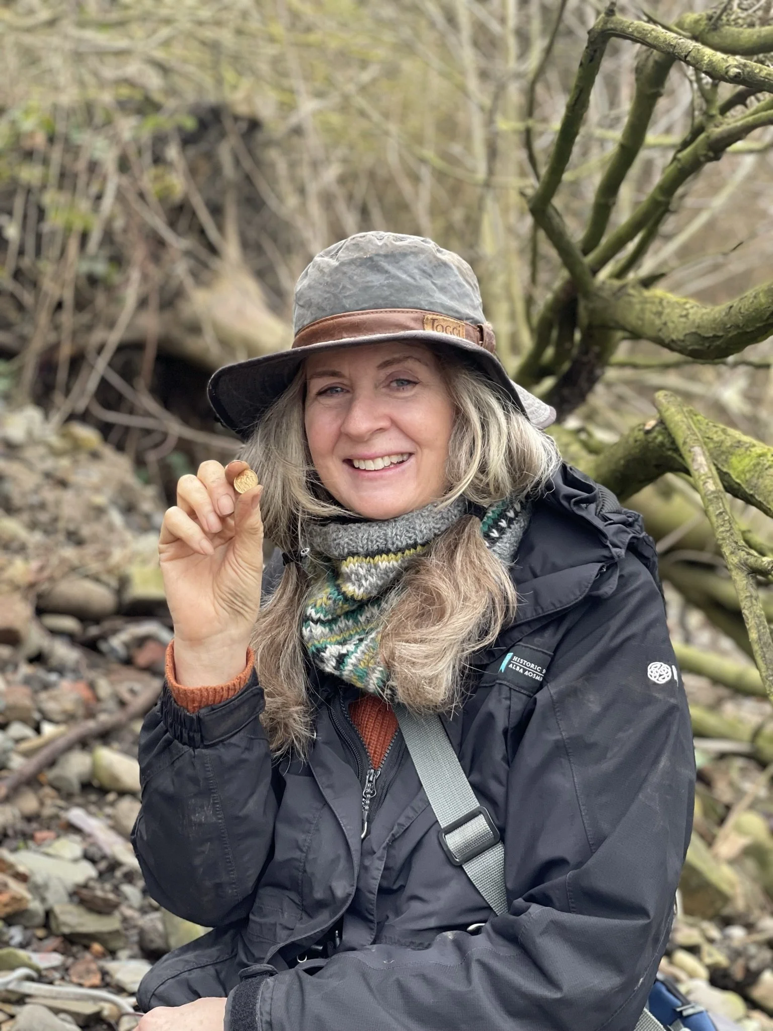 A smiling woman wearing a wide-brimmed hat, a scarf, and a waterproof jacket, sitting on a river foreshore, holding a gold coin