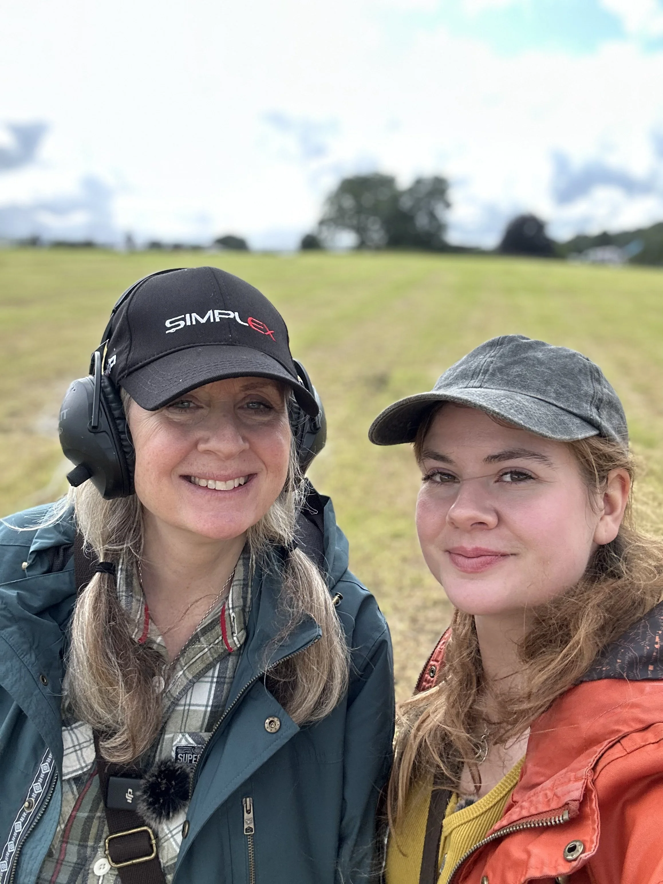 Two women outdoors in a grassy field, smiling for a selfie. One is wearing a black cap, headphones, and a blue jacket. The other is wearing a gray cap and a colorful jacket.