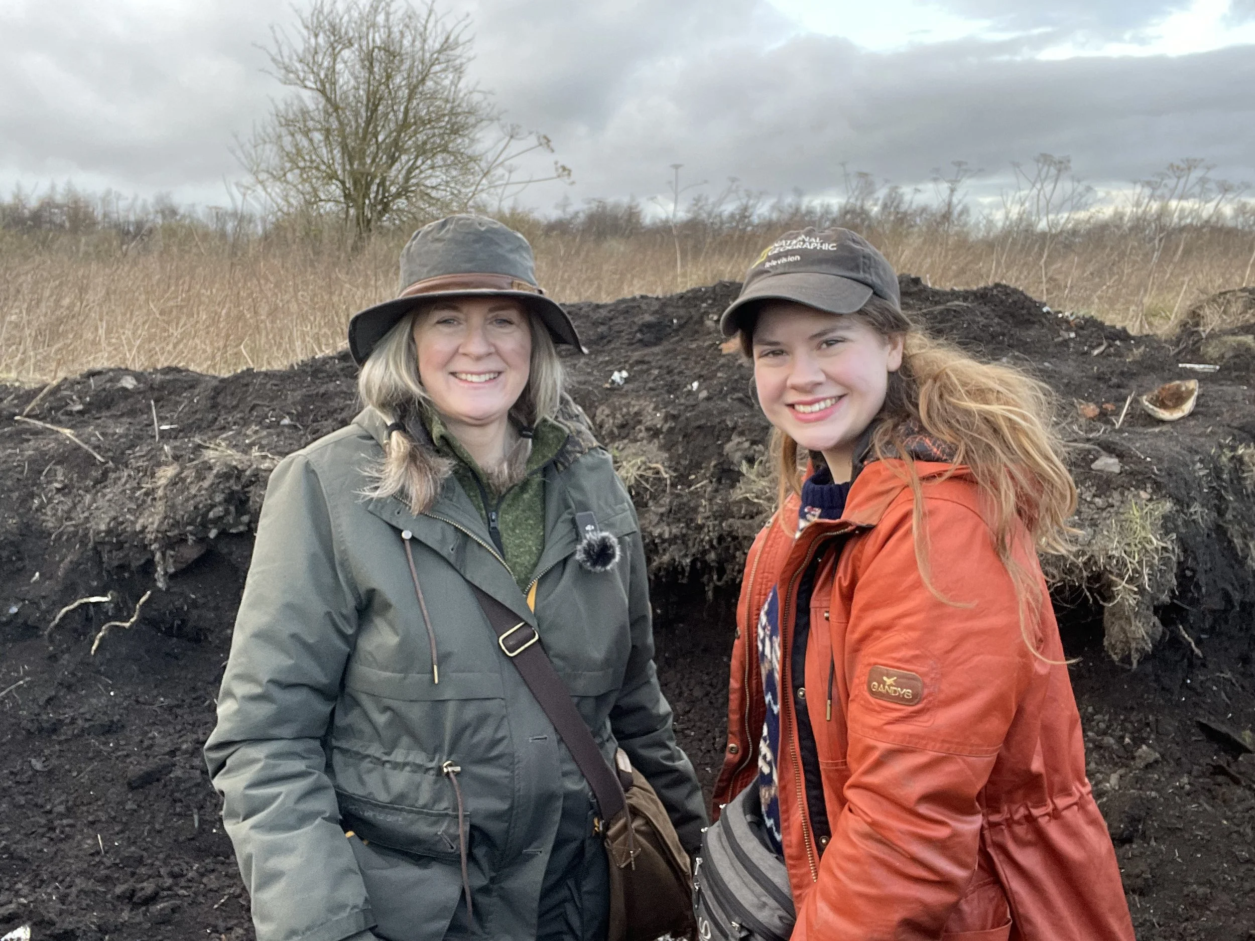 Two women outdoors in a bottle dump standing in front of a large mound of dirt. One woman is wearing a green jacket and a hat, and the other is in an orange jacket and a cap. Both are smiling.