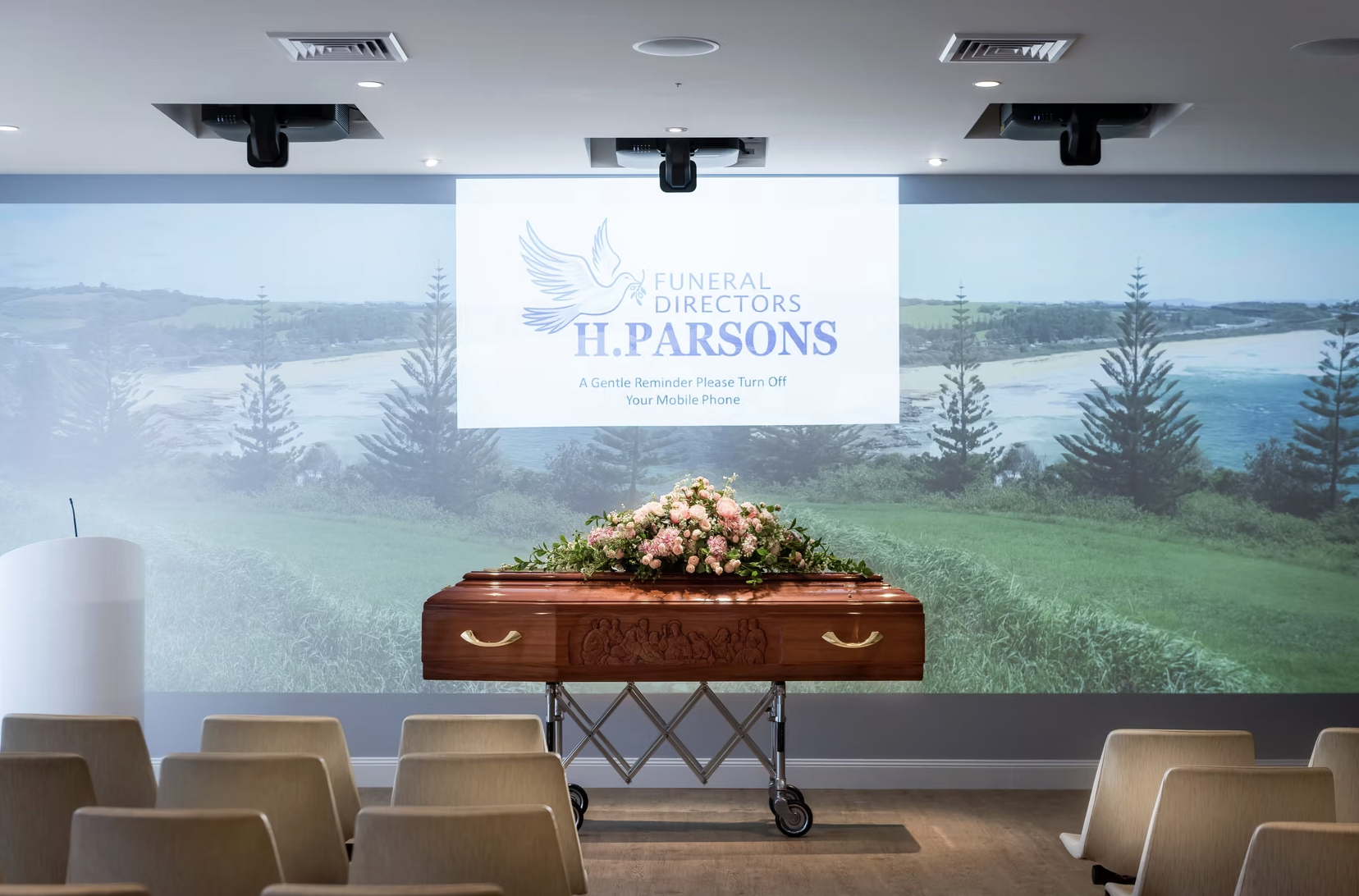 Funeral home setting with a closed casket decorated with pink and white flowers, chairs arranged for a funeral service, and a projection on the wall displaying the name H. Parsons Funeral Directors with a dove logo and a reminder to turn off mobile p