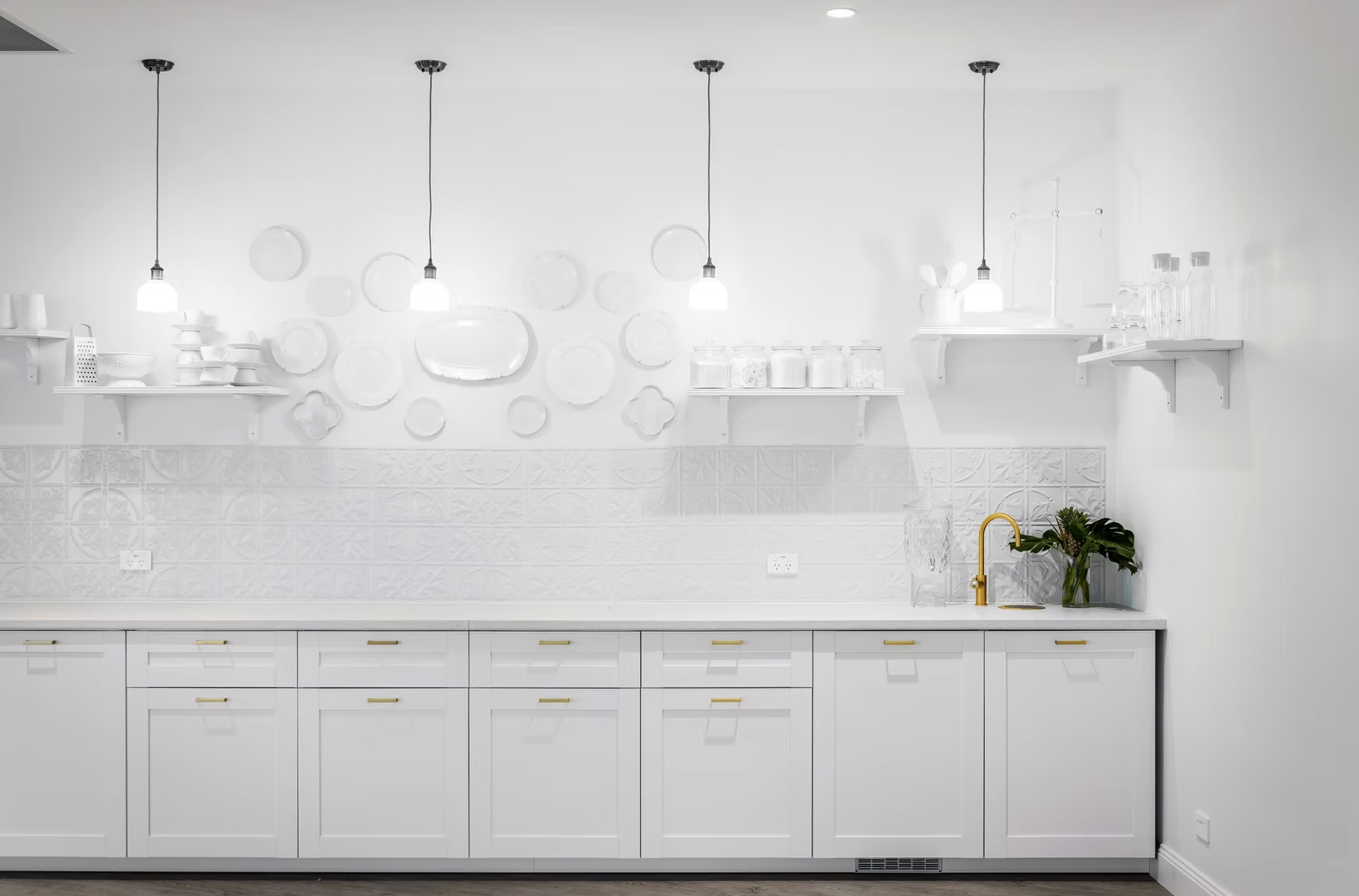 Modern white kitchen with gold faucet and open shelving, decorated with plants, jars, and dishes.