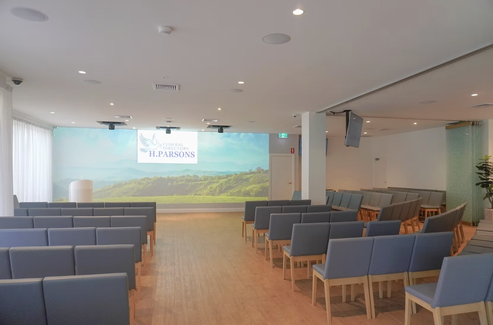 Empty funeral home chapel with rows of blue chairs facing a large screen displaying a logo and text for 'Funeral Directors H. Parsons.' The room has natural light from windows on the left, light-colored walls, a wood floor, and ceiling-mounted lights