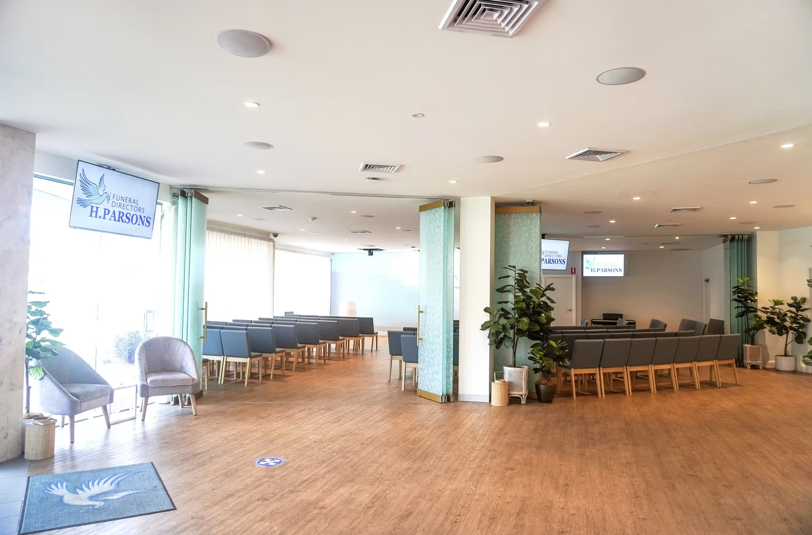 Empty funeral home waiting room with chairs, potted plants, and multiple screens displaying 'Funeral Directors H. Parson.'