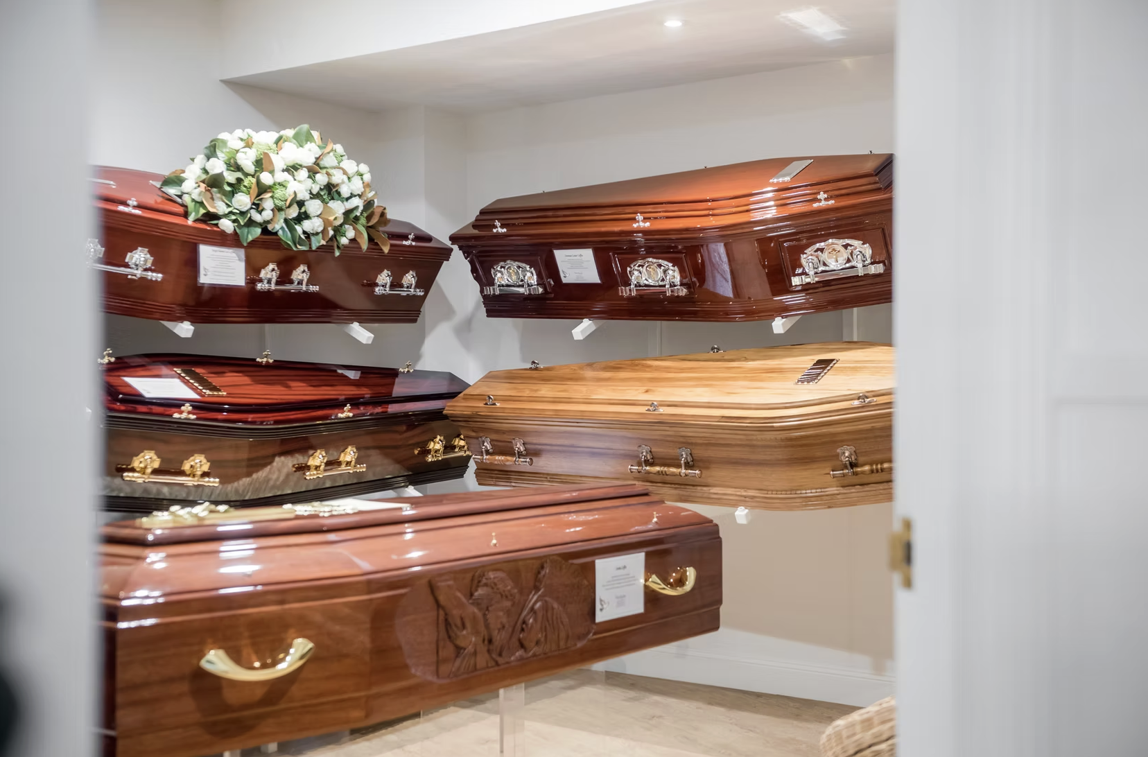 Display of various wooden coffins with different finishes and designs, arranged on shelves in a showroom, with a floral arrangement on one of the coffins.