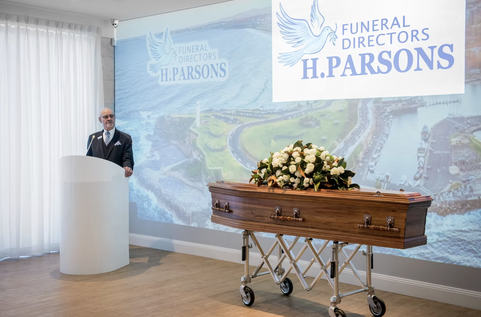 A man in a dark suit standing at a white podium during a funeral service. There is a wooden casket with a floral arrangement on top in front of him. A large screen behind displays a logo for funeral directors H. Parsons with a dove and water backgrou