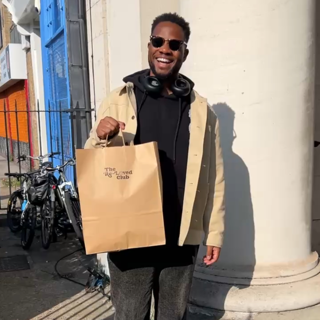 A smiling man with sunglasses holding a shopping bag in front of a white column, with bicycles and a colorful storefront in the background.