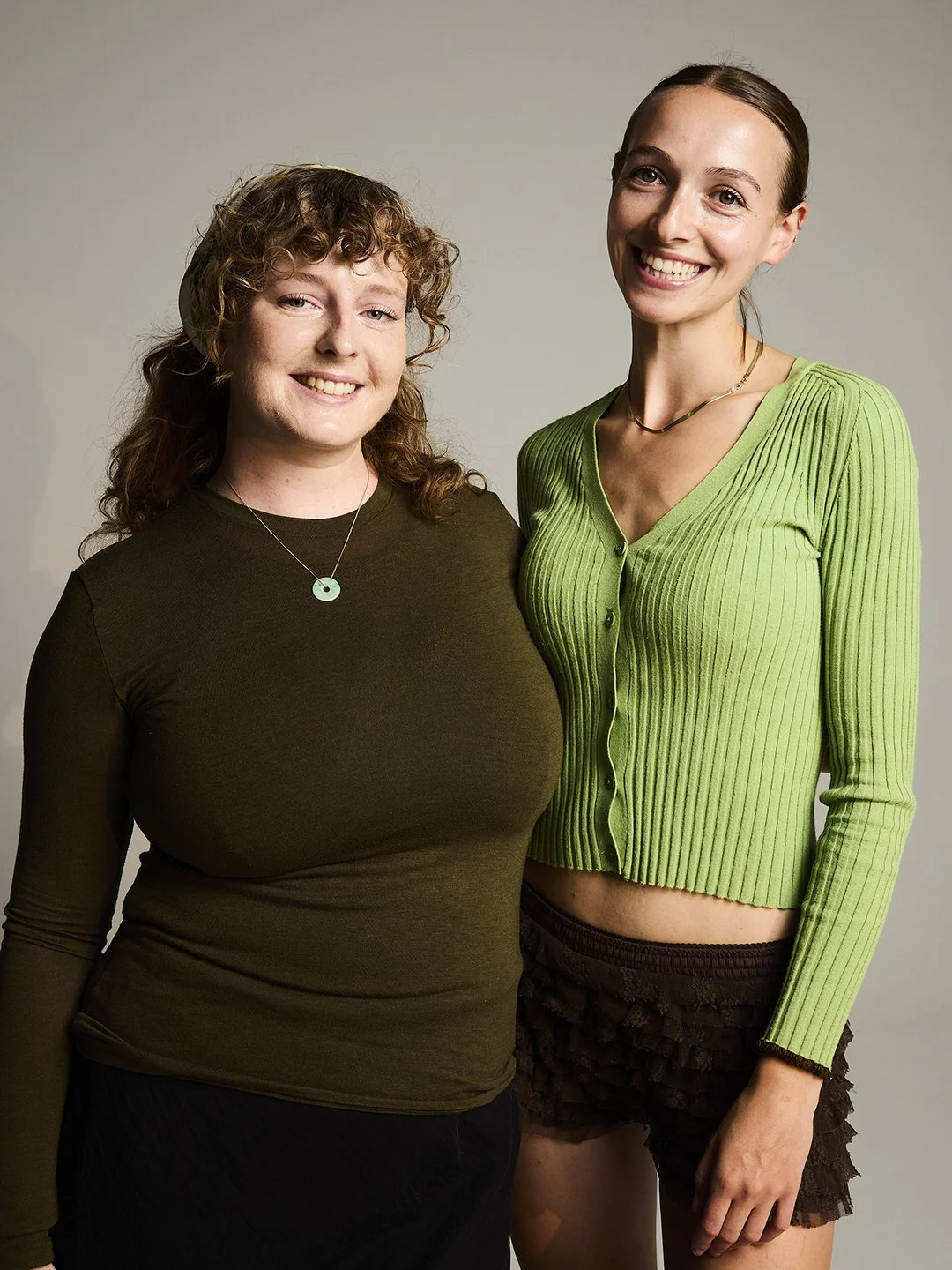 Two friends standing side by side, smiling, against a plain background. One is wearing a dark green long-sleeve shirt with a necklace, and the other is in a bright green ribbed cardigan with a necklace and shorts. Sellers at The Re-Loved Club.